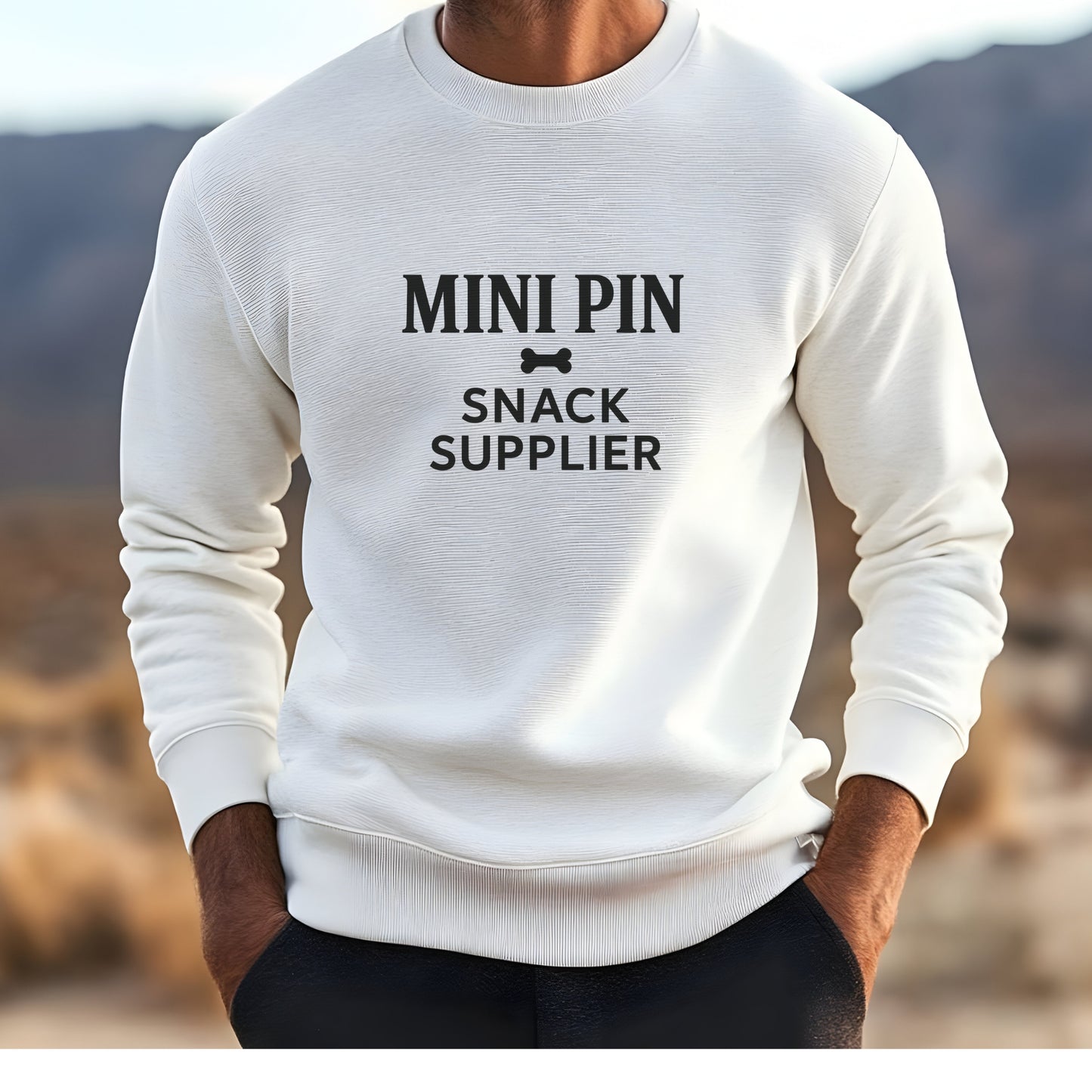 Man wearing a white "Mini Pin Snack Supplier" Miniature Pinscher unisex sweatshirt outdoors in the mountains