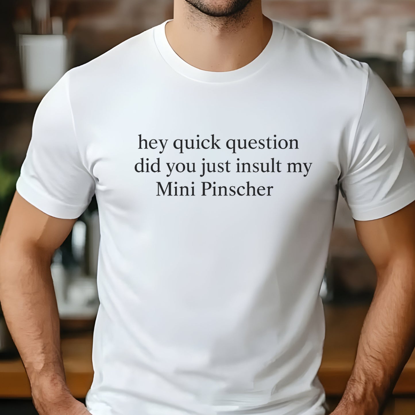 Man wearing a white "hey quick question did you just insult my Mini Pinscher" t-shirt, standing by a window