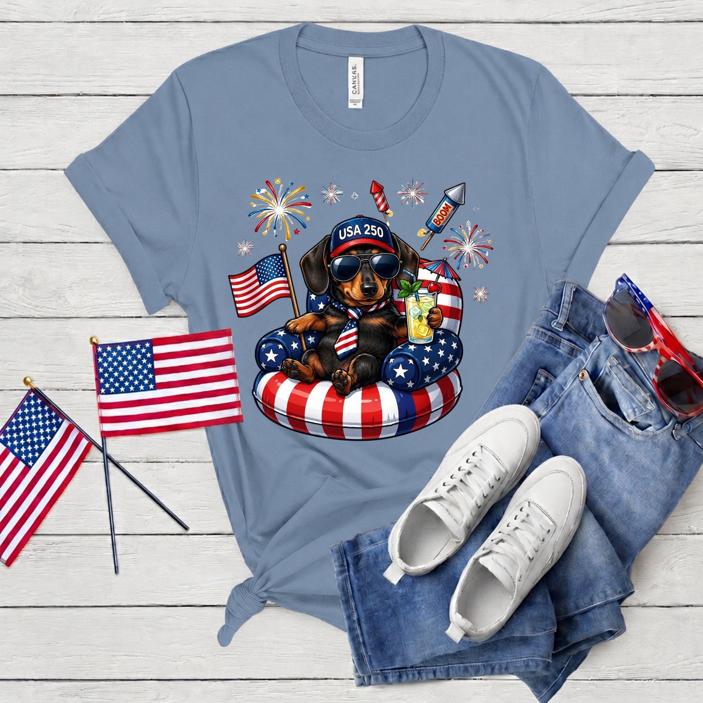 Blue t-shirt with a dachshund graphic, American flags, and sunglasses on a wooden surface.