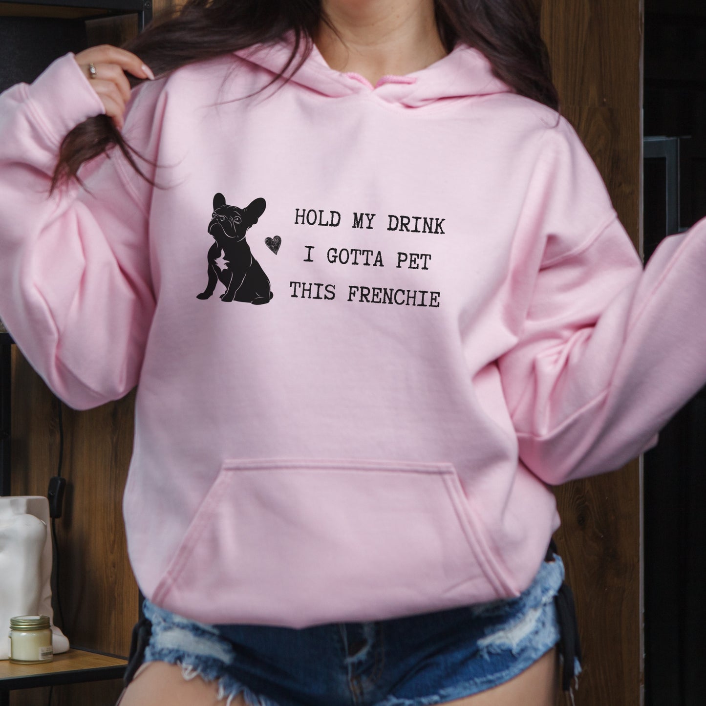 Light pink hoodie with a black line-art French Bulldog illustration and the bold text “Hold my drink, I gotta pet this Frenchie” printed on the front.