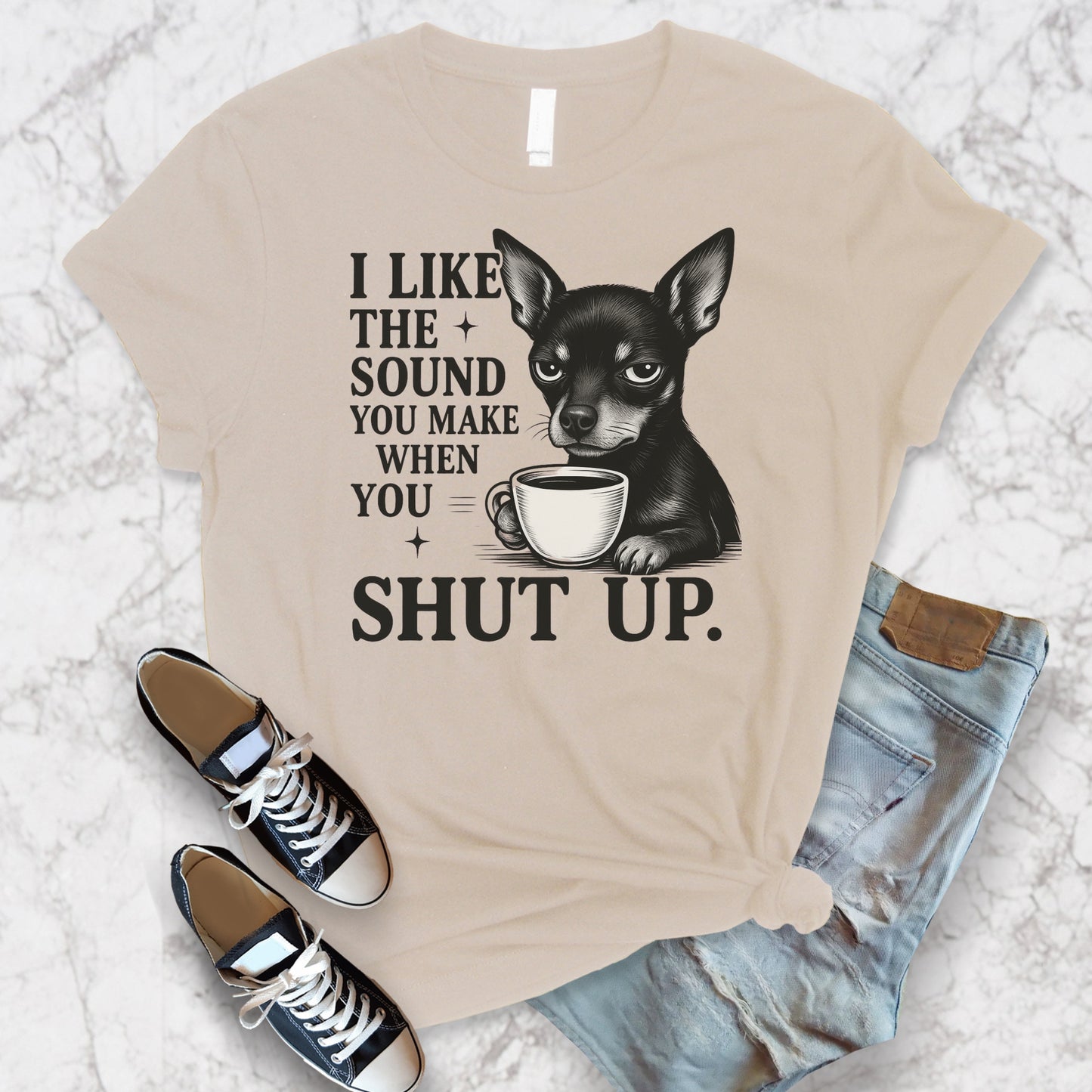 Flay lay of a soft cream "I Like The Sound You Make When You Shut Up" Mini Pinscher unisex t-shirt holding coffee
