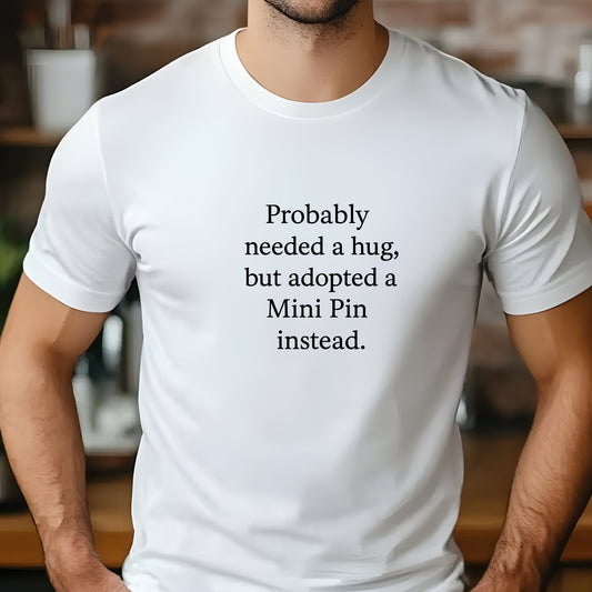 White unisex t-shirt with white text: "Probably needed a hug, but adopted a Mini Pin instead." displayed on plain background 