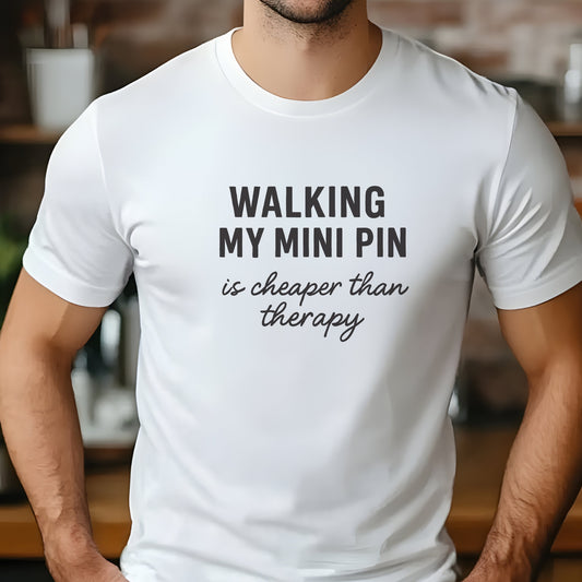 White unisex t-shirt with black text: "WALKING MY MINI PIN is cheaper than therapy" centered on chest