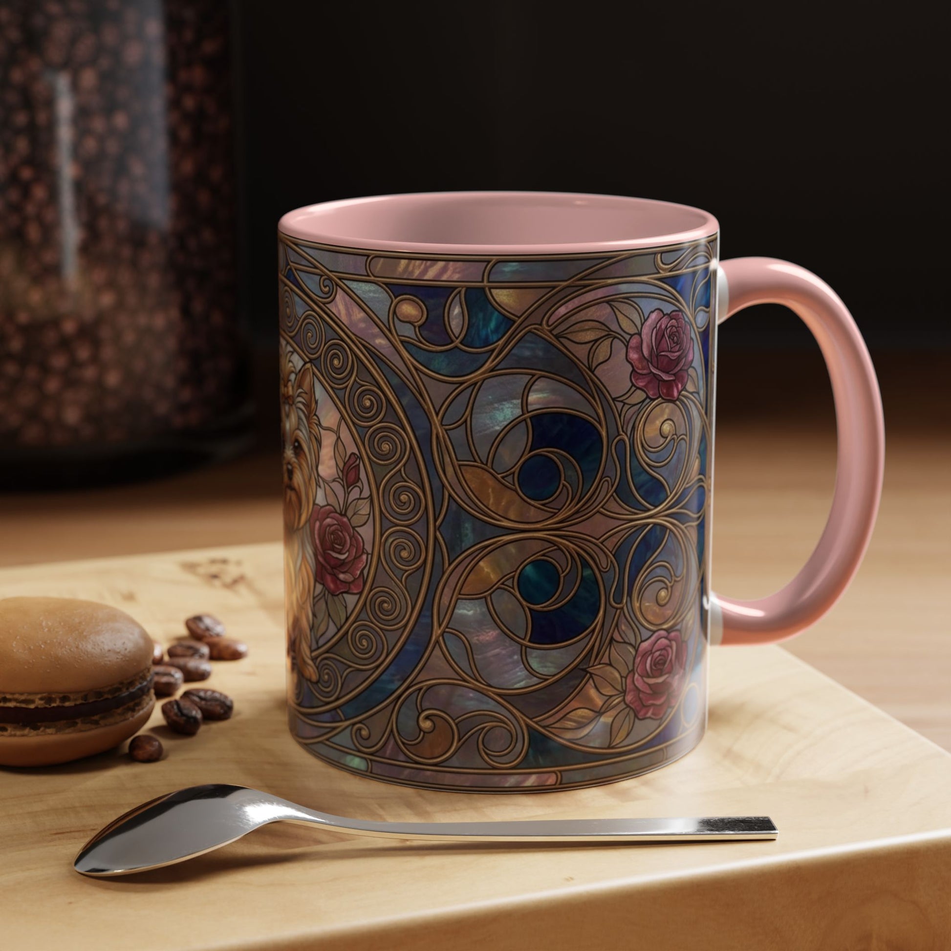 A pink ceramic mug with an ornate stained-glass-style floral pattern in blue, gold, and rose tones, shown on a wooden surface with a spoon and coffee beans nearby.