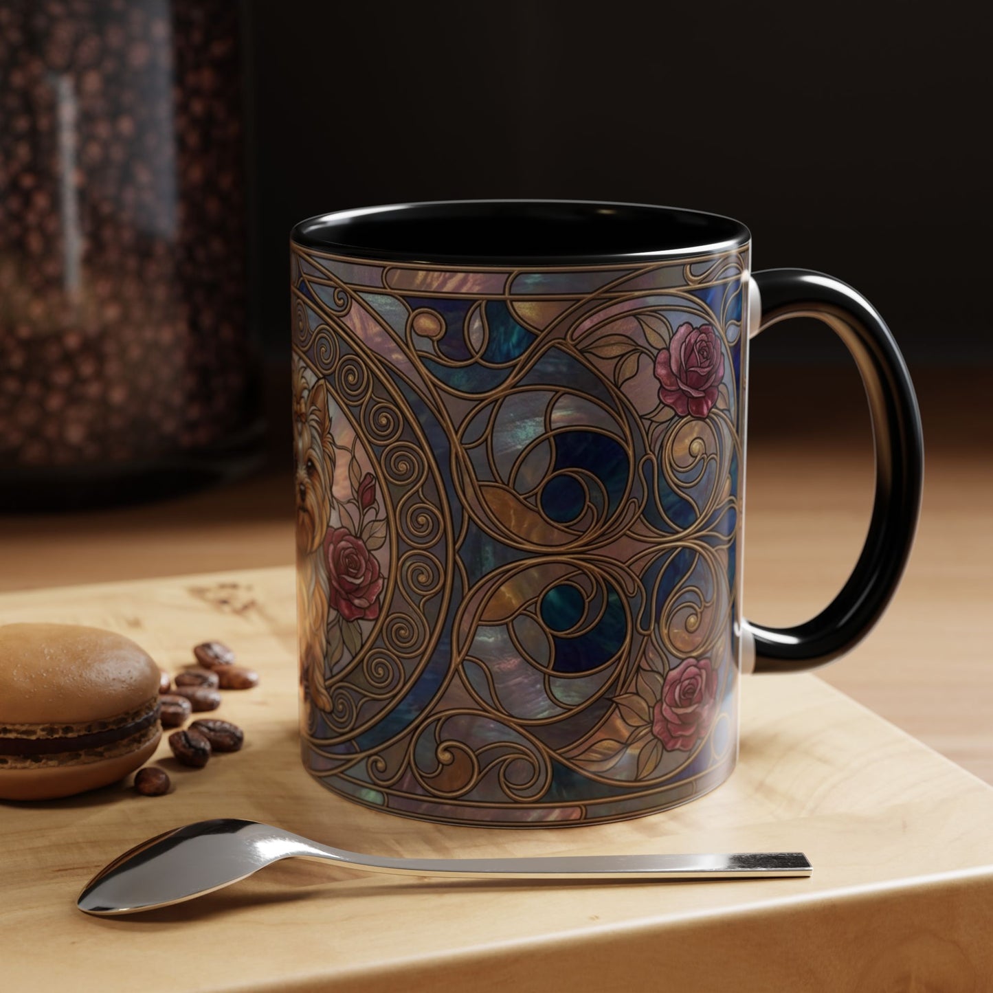A ceramic mug with a black handle and interior, decorated with an ornate Art Nouveau stained-glass style pattern in blue, gold, and pink. The mug is set on a light wooden surface next to a spoon, with a blurred background showing coffee beans and a macaron.