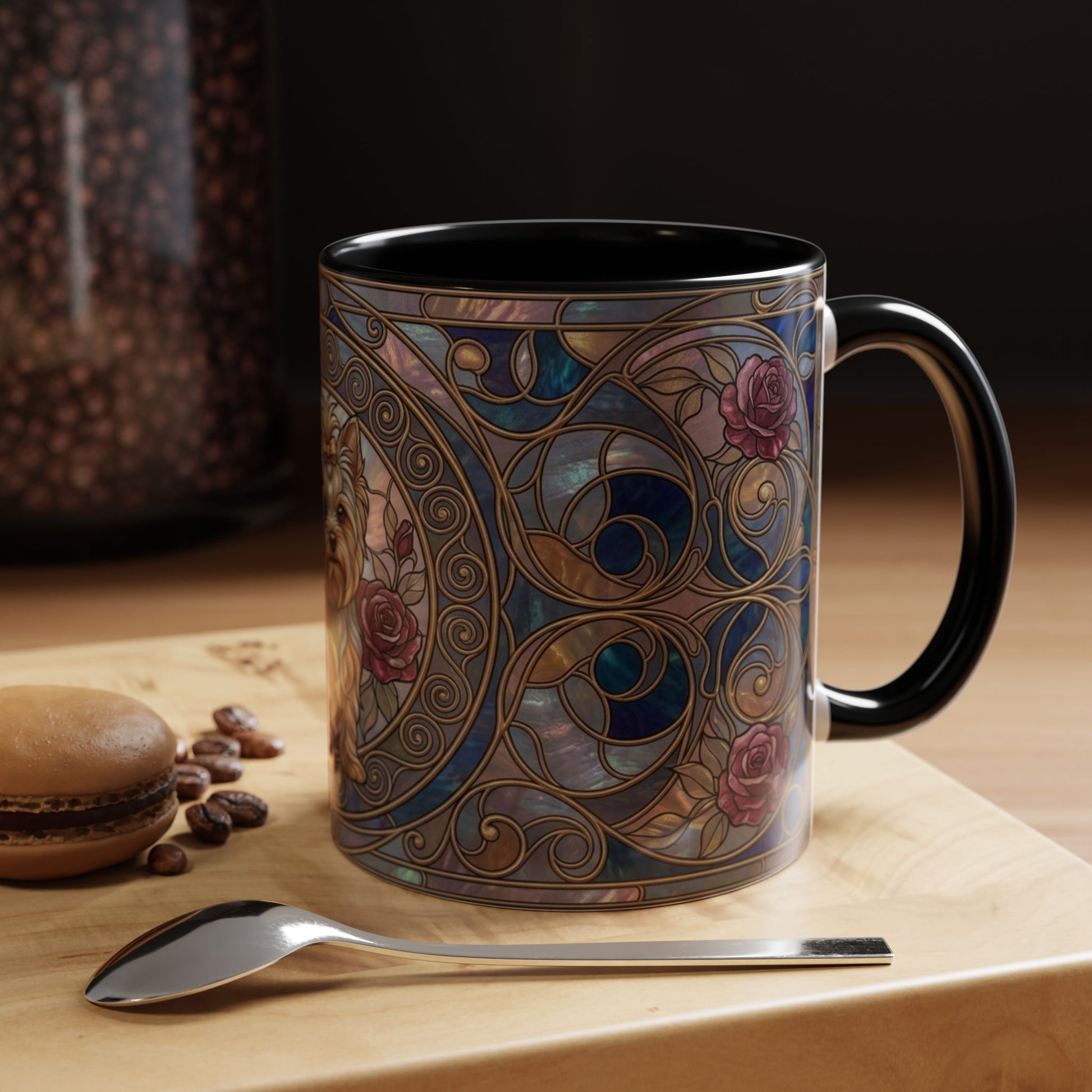 A ceramic mug with a black handle and interior, decorated with an ornate Art Nouveau stained-glass style pattern in blue, gold, and pink. The mug is set on a light wooden surface next to a spoon, with a blurred background showing coffee beans and a macaron.