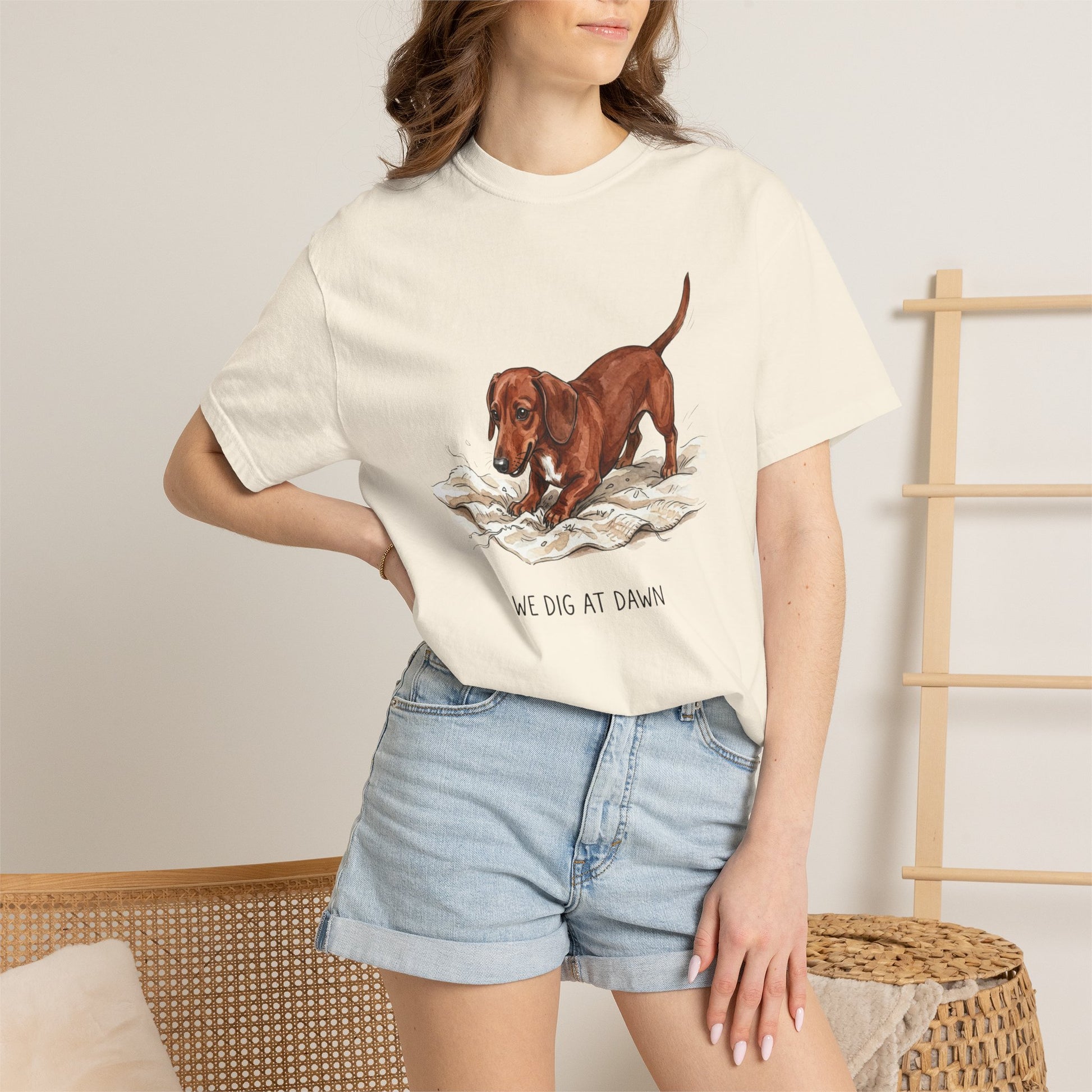 A woman wearing a cream-colored graphic t-shirt with a brown dachshund illustration and the text 'WE DIG AT DAWN', paired with light blue denim shorts in a warm, neutral-toned room setting.