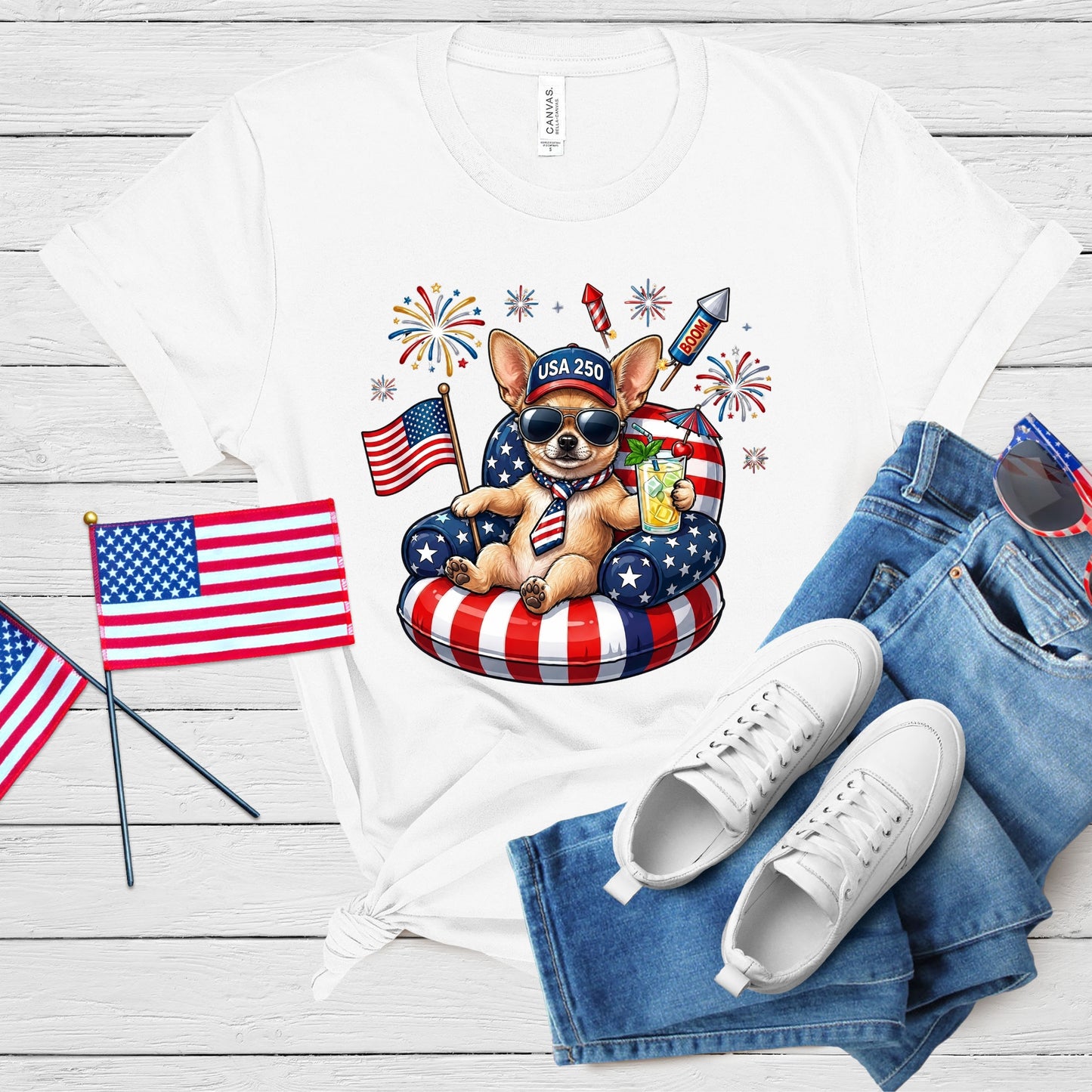 White t-shirt with a dog graphic, American flags, and sunglasses on a wooden surface.