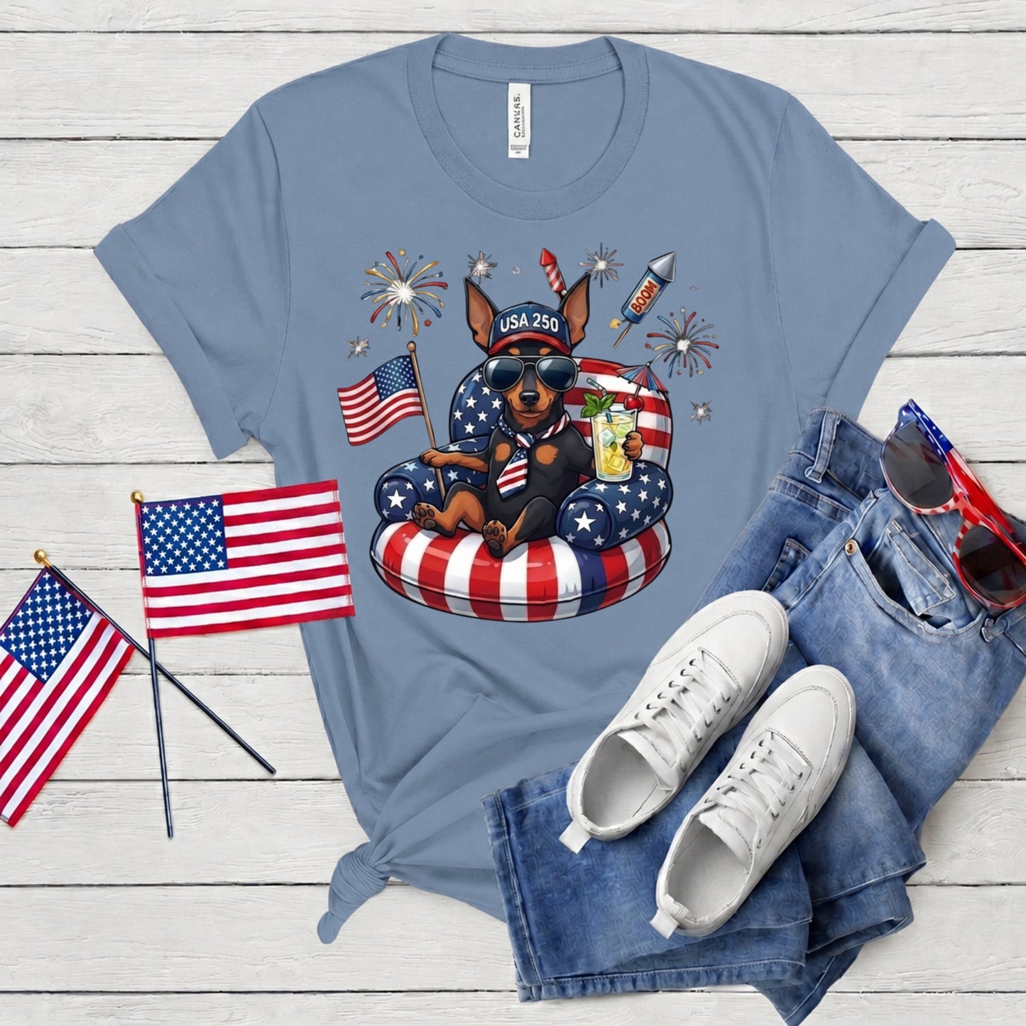Blue t-shirt with a dog graphic, American flags, and sunglasses on a wooden surface.