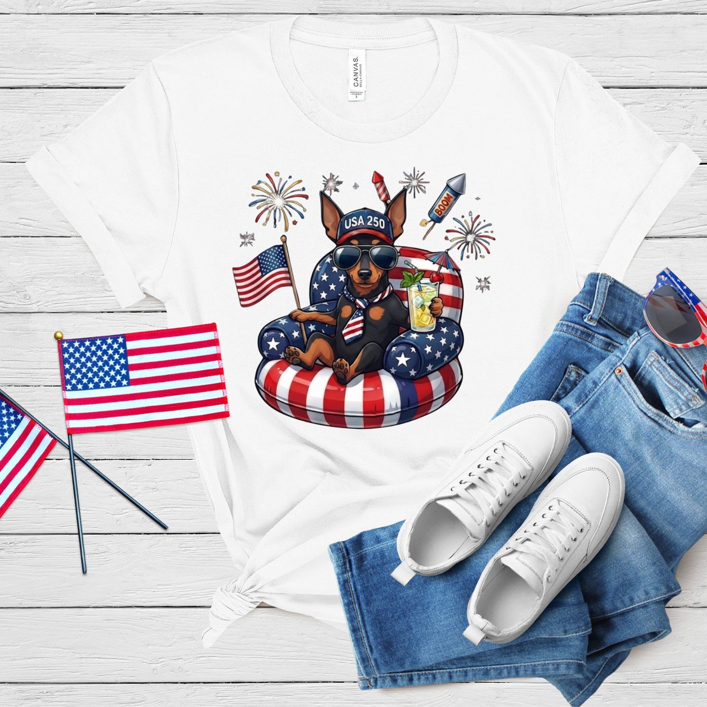 White t-shirt with a dog graphic, American flags, and sunglasses on a wooden surface.