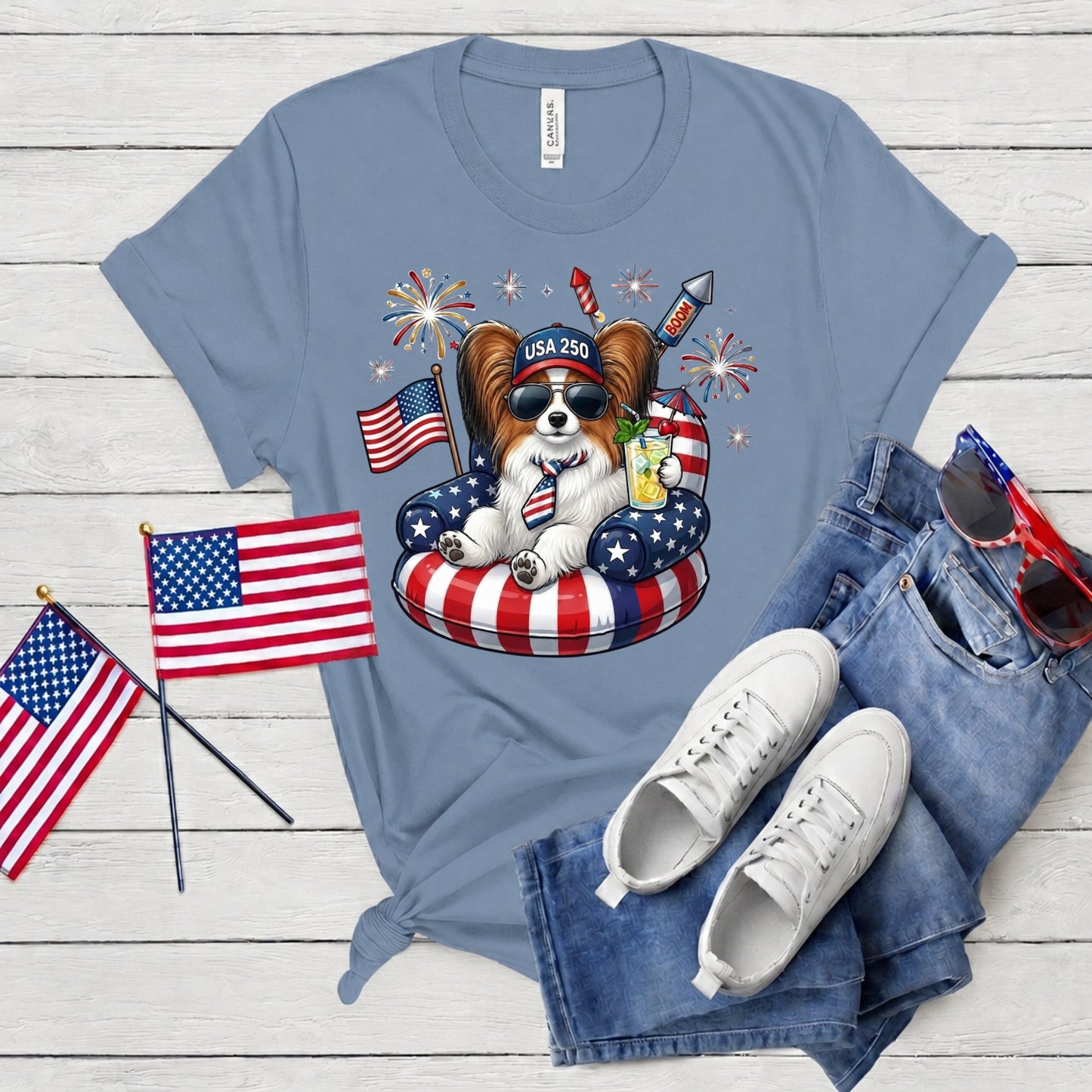 Blue t-shirt with a dog graphic, American flags, and sunglasses on a wooden surface.