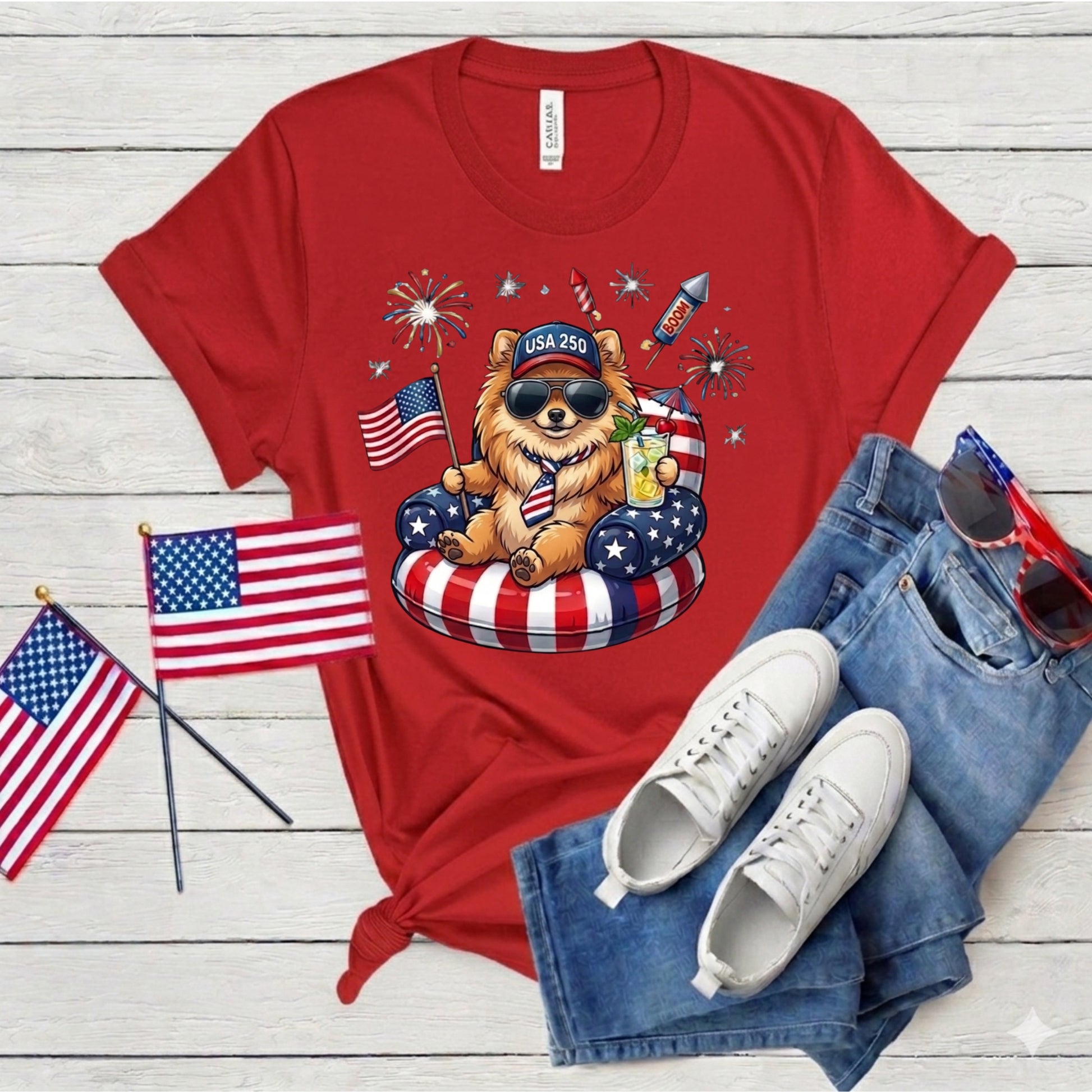 Red t-shirt with a bear graphic, sunglasses, and American flags on a wooden surface with white sneakers and blue jeans.