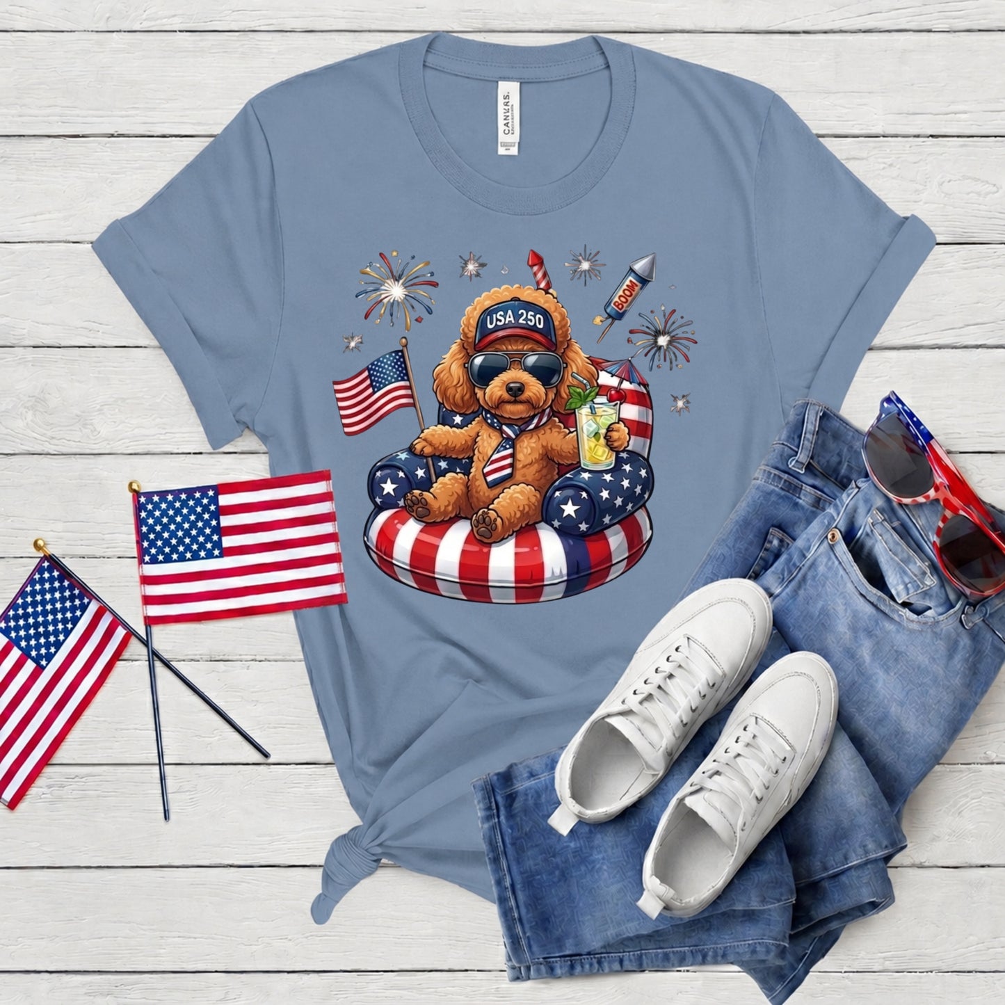 Blue t-shirt with a teddy bear graphic, American flags, and sunglasses on a wooden surface.