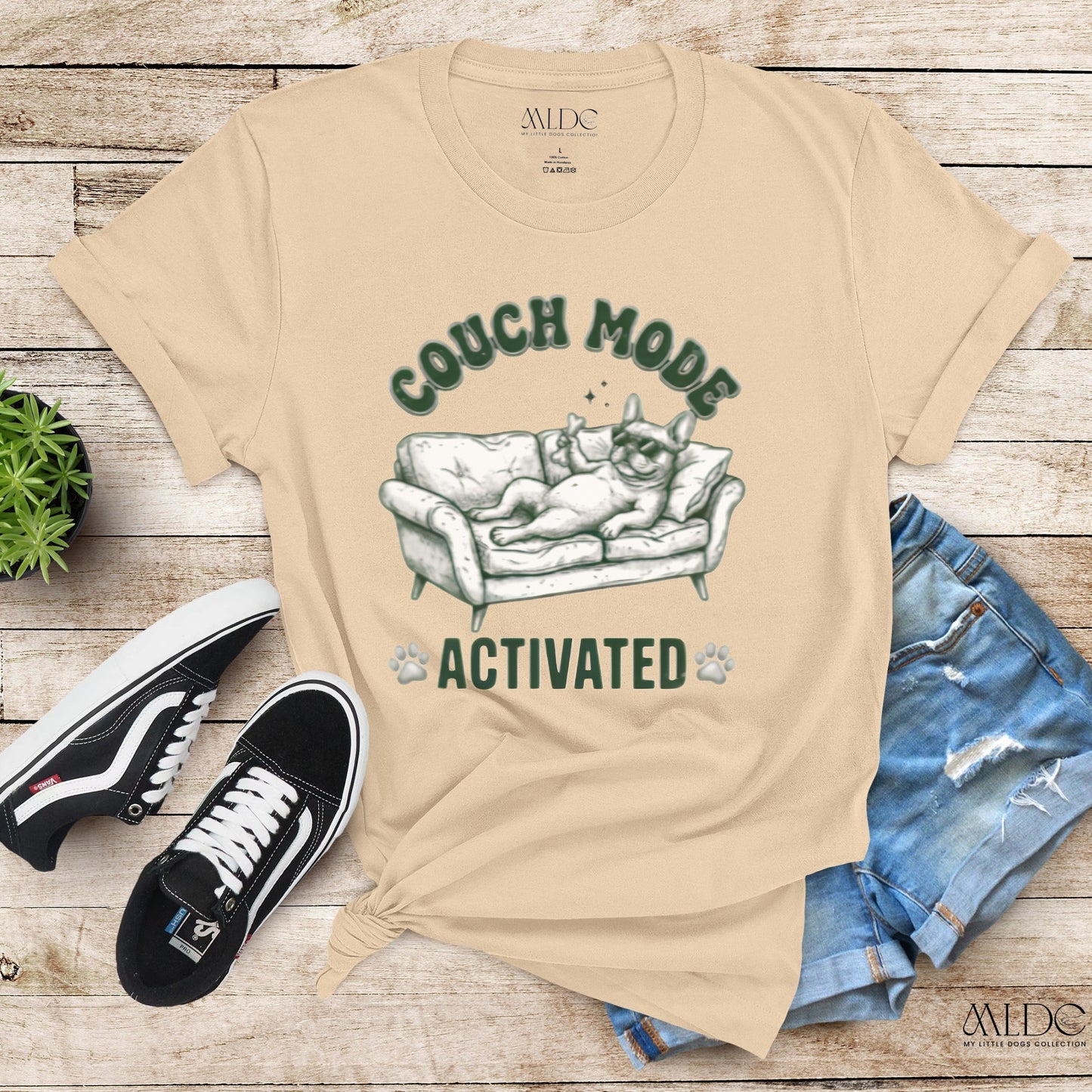 Beige t-shirt with 'Couch Mode Activated' graphic on a wooden surface with sneakers and jeans.
