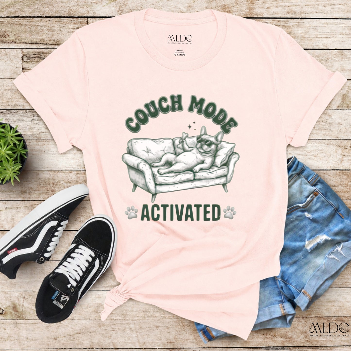 Pink t-shirt with 'Couch Mode Activated' graphic, black sneakers, and jeans on a wooden surface.