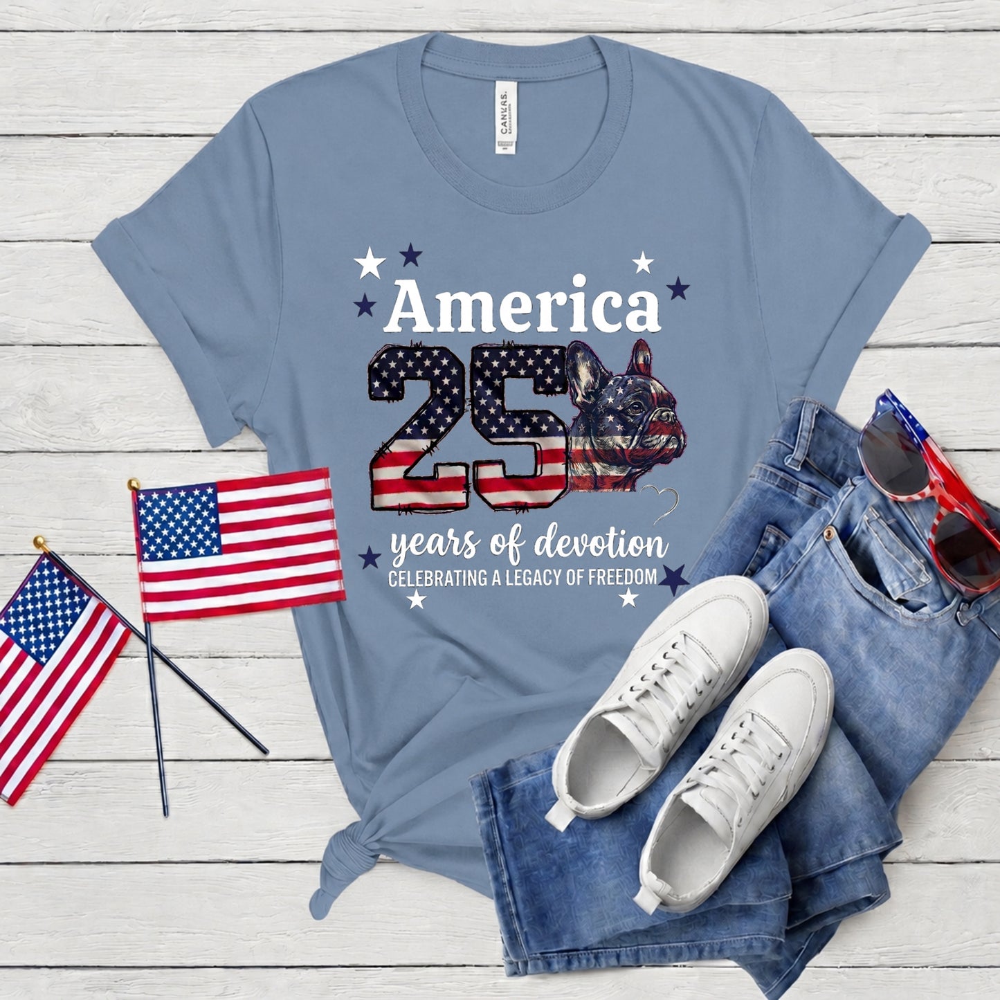 Blue t-shirt with American-themed design, sunglasses, jeans, and flags on a wooden surface