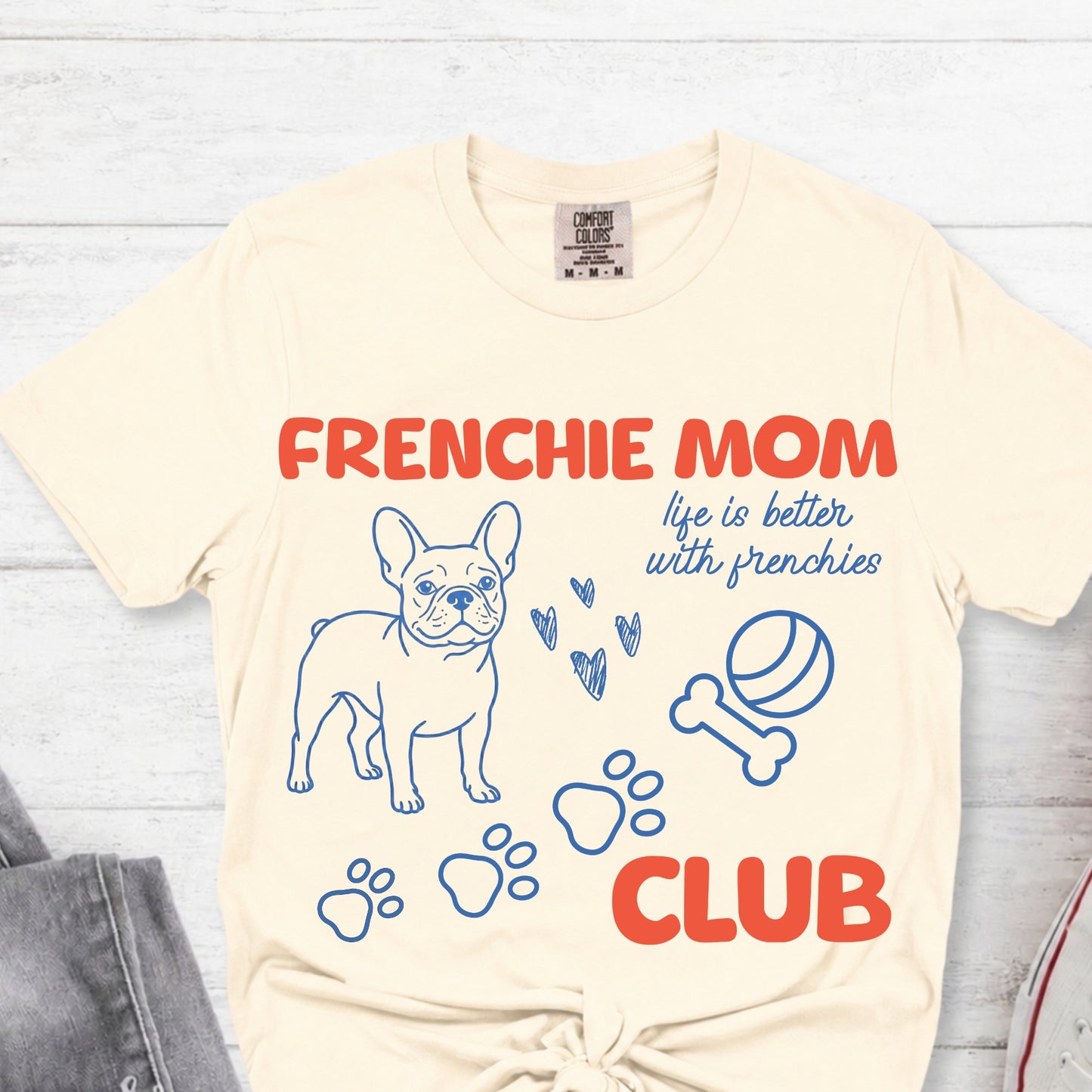 Beige t-shirt with 'Frenchie Mom Club' text and graphics on a light wooden surface.