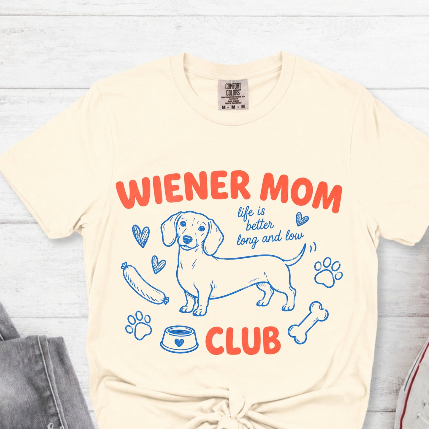 Beige t-shirt with 'Wiener Mom Club' graphic design on a light wooden surface.