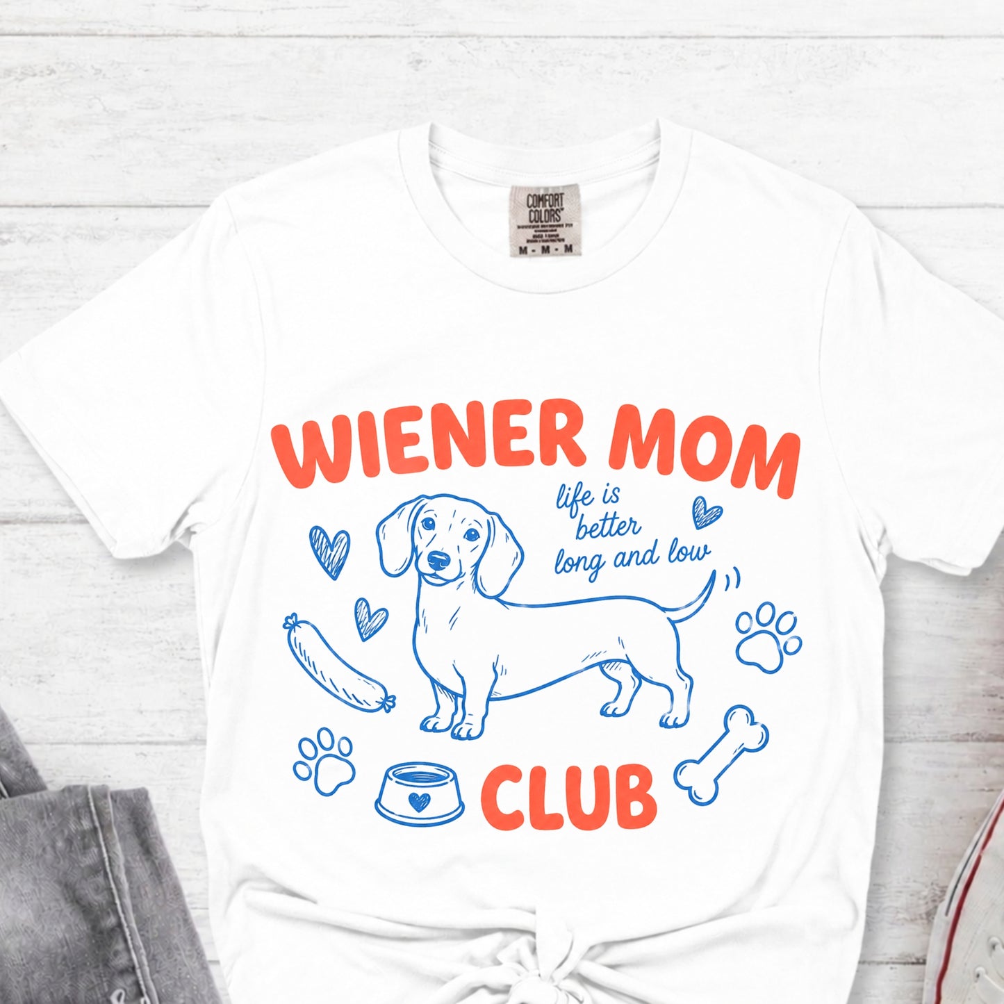 White t-shirt with 'Wiener Mom Club' graphic design on a light wooden surface.