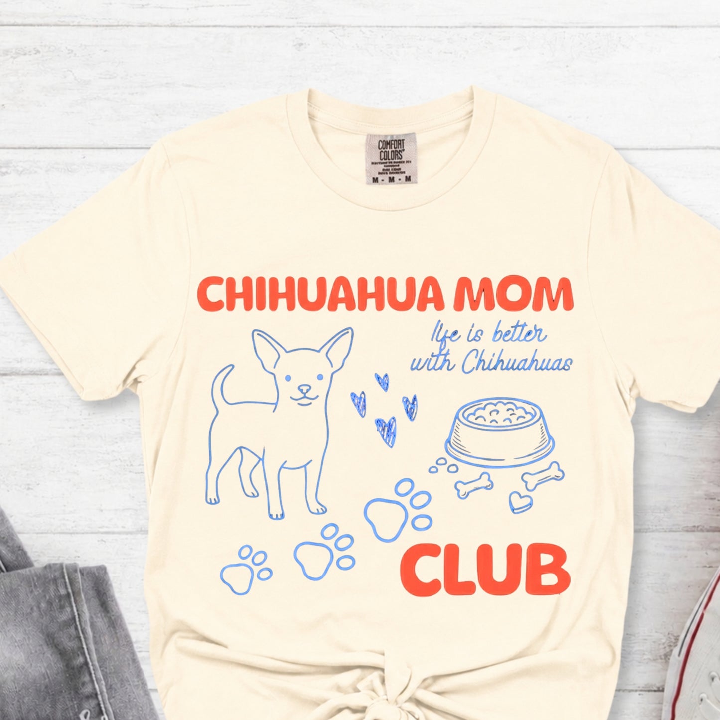 Beige t-shirt with 'Chihuahua Mom Club' text and graphics on a light wooden surface.