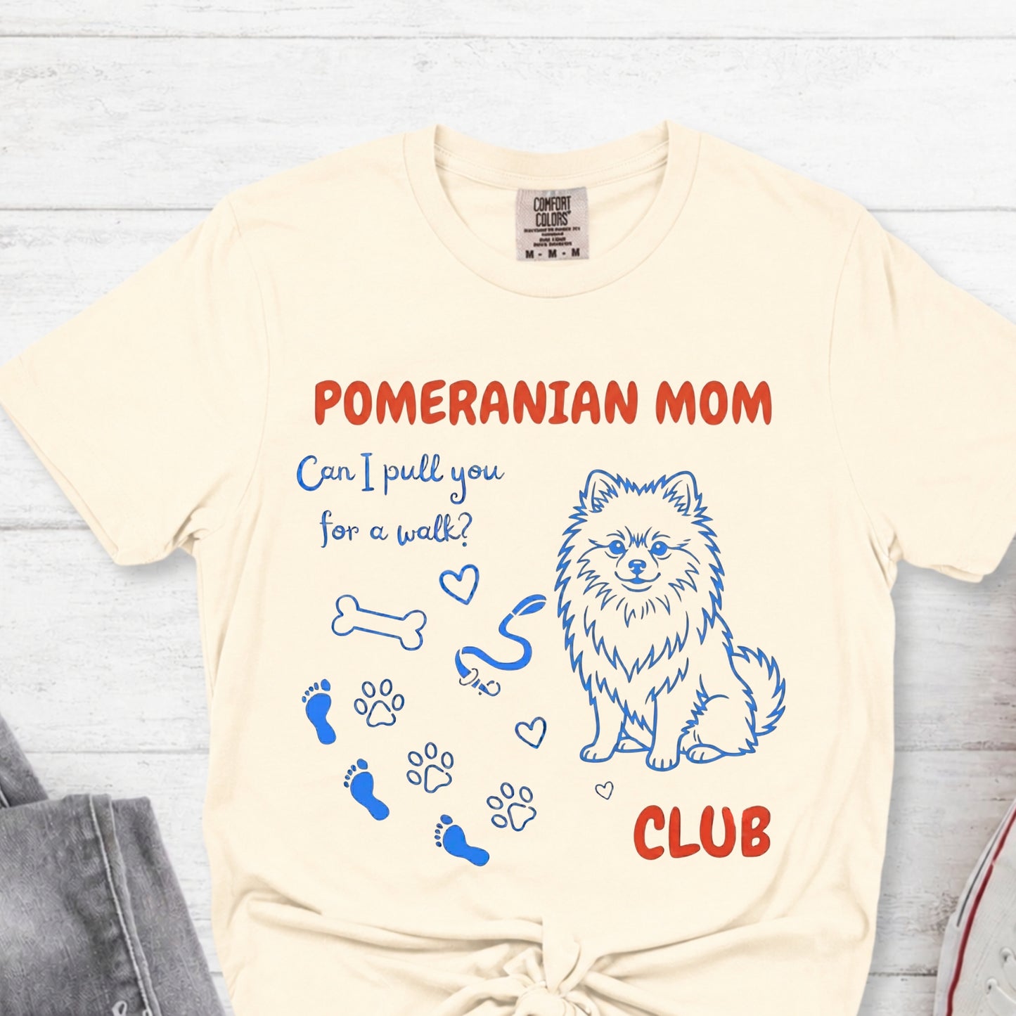 Beige t-shirt with Pomeranian-themed design and text on a light wooden surface.