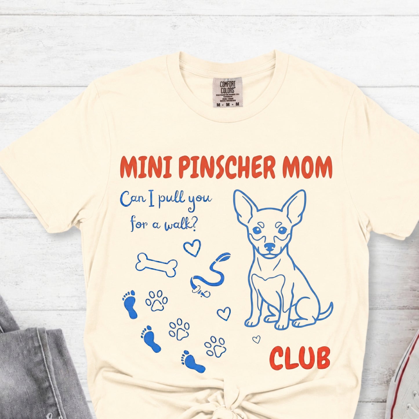 Beige t-shirt with 'Mini Pinscher Mom' design on a light wooden surface