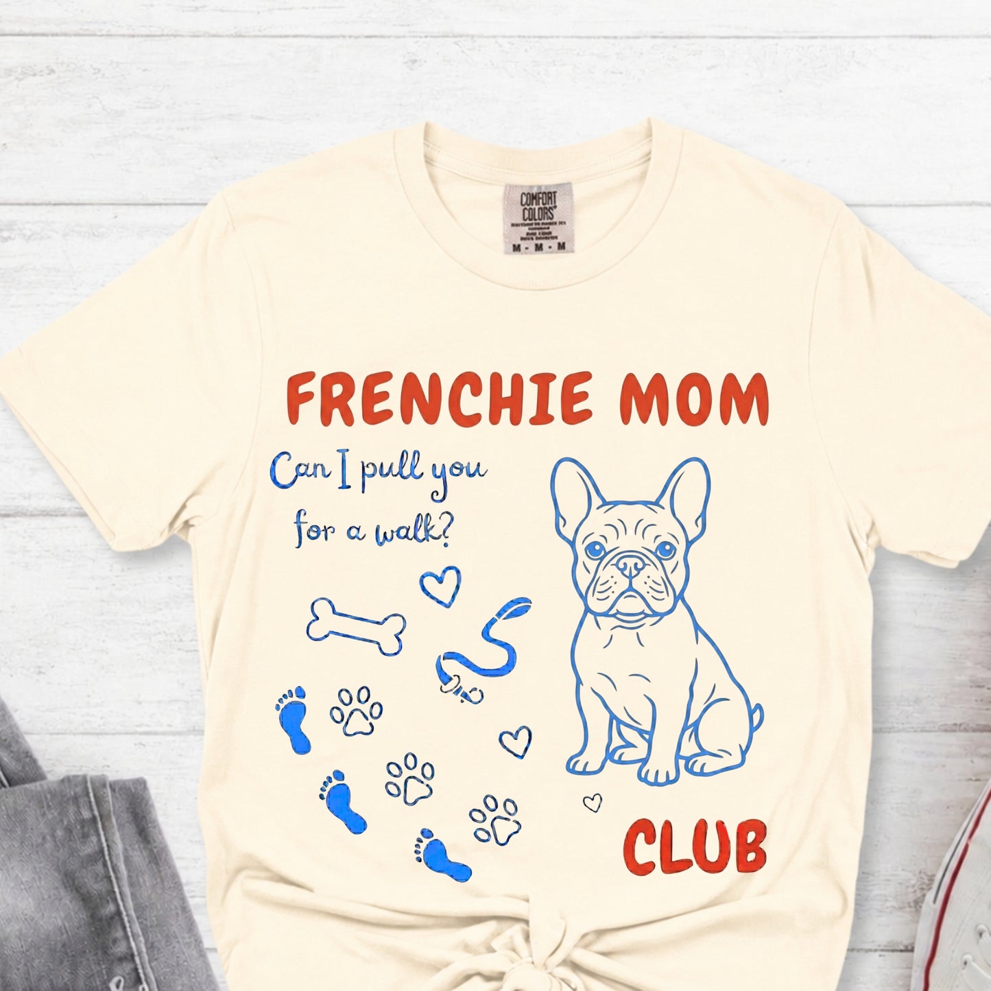 Beige t-shirt with Frenchie Mom graphic design on a light wooden surface