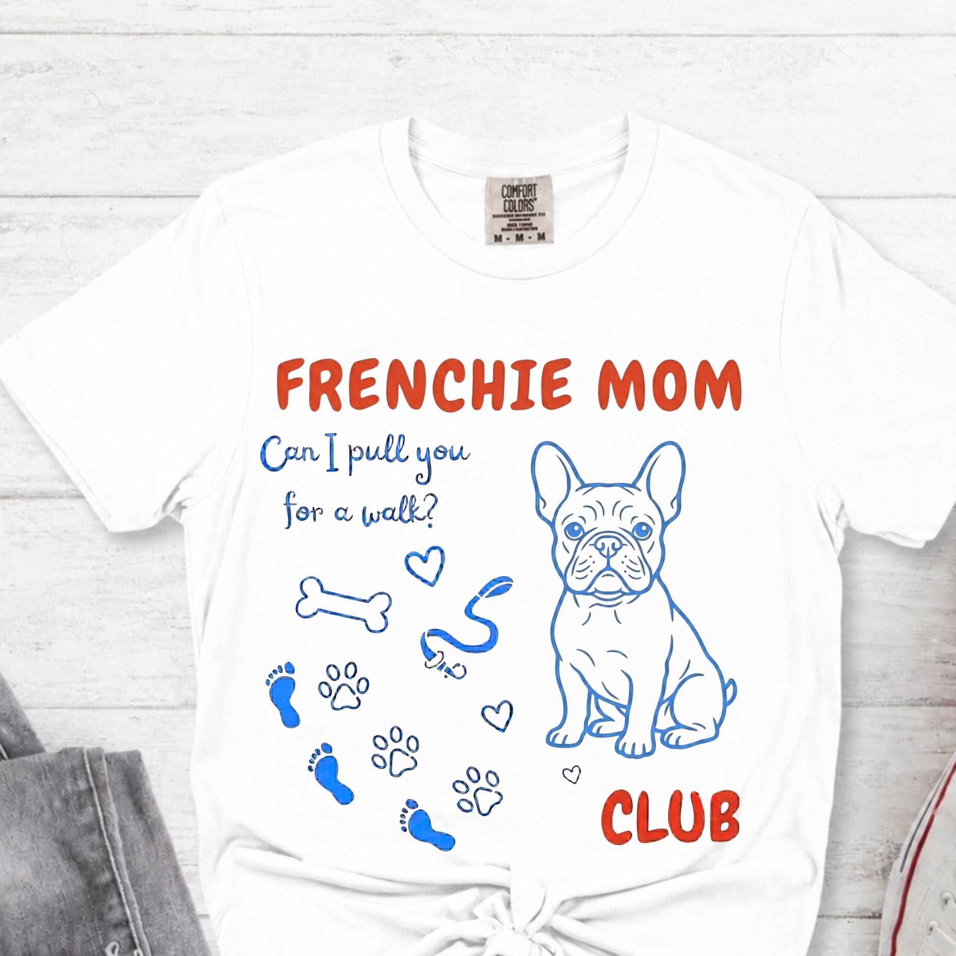 White t-shirt with 'FRENCHIE MOM' text and dog illustrations on a light wooden surface.