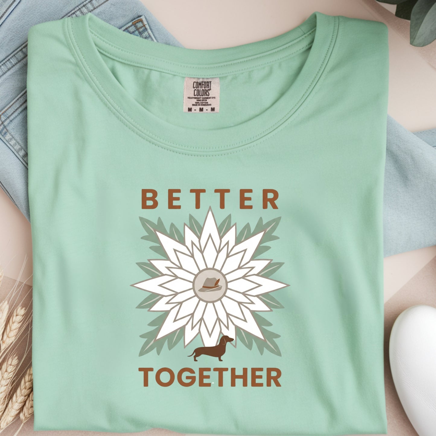Folded light green t-shirt with a flower design and 'Better Together' text on a neutral background.