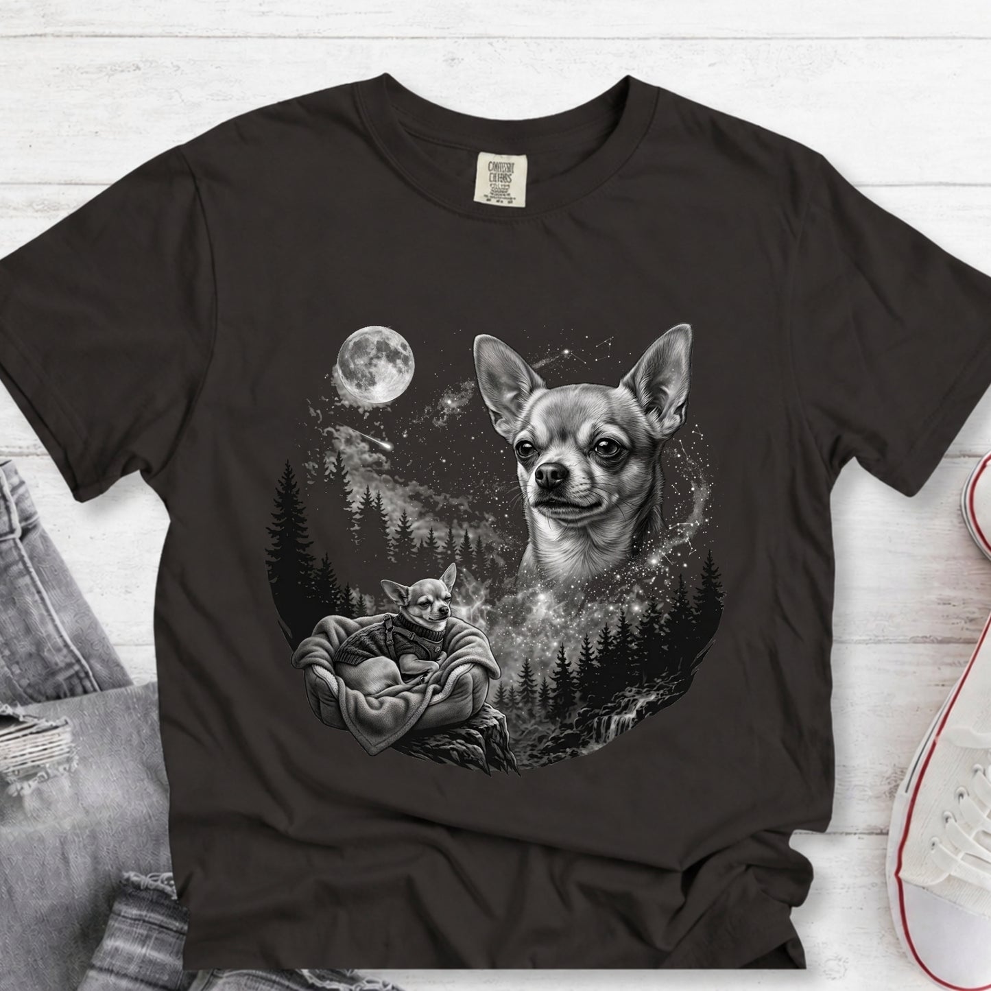 A black short-sleeve graphic T-shirt laid flat on a white wood surface, featuring a grayscale print of a Chihuahua moon dog and a Star Wars Yoda figure. Partial jeans and a white sneaker are visible at the edges as styling props.