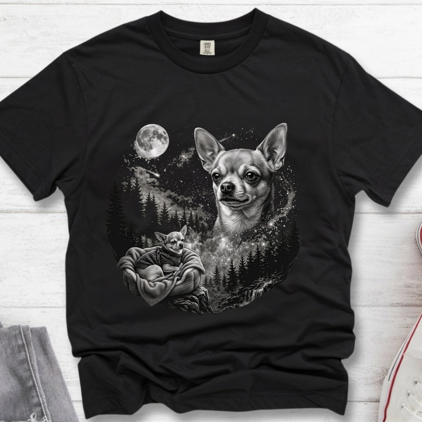 A black short-sleeve crewneck t-shirt laid flat on a white wood background, featuring a detailed black and white graphic print of a Chihuahua dog and a sleeping cat under a full moon in a forest.