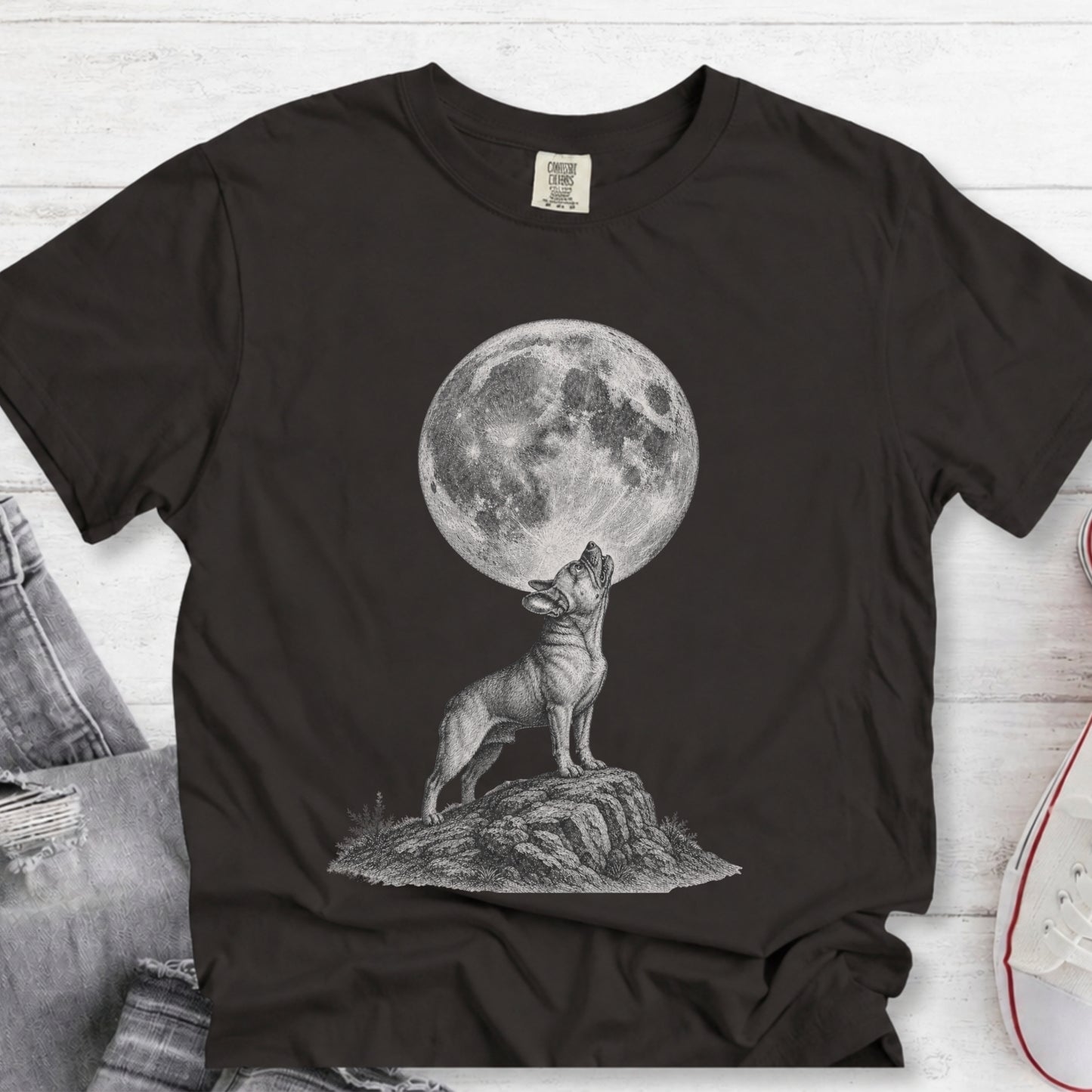 A black short-sleeve graphic T-shirt laid flat on a white wood background, featuring a monochrome illustration of a dog howling at a large full moon. Jeans and part of a shoe are visible as styling props around the shirt.