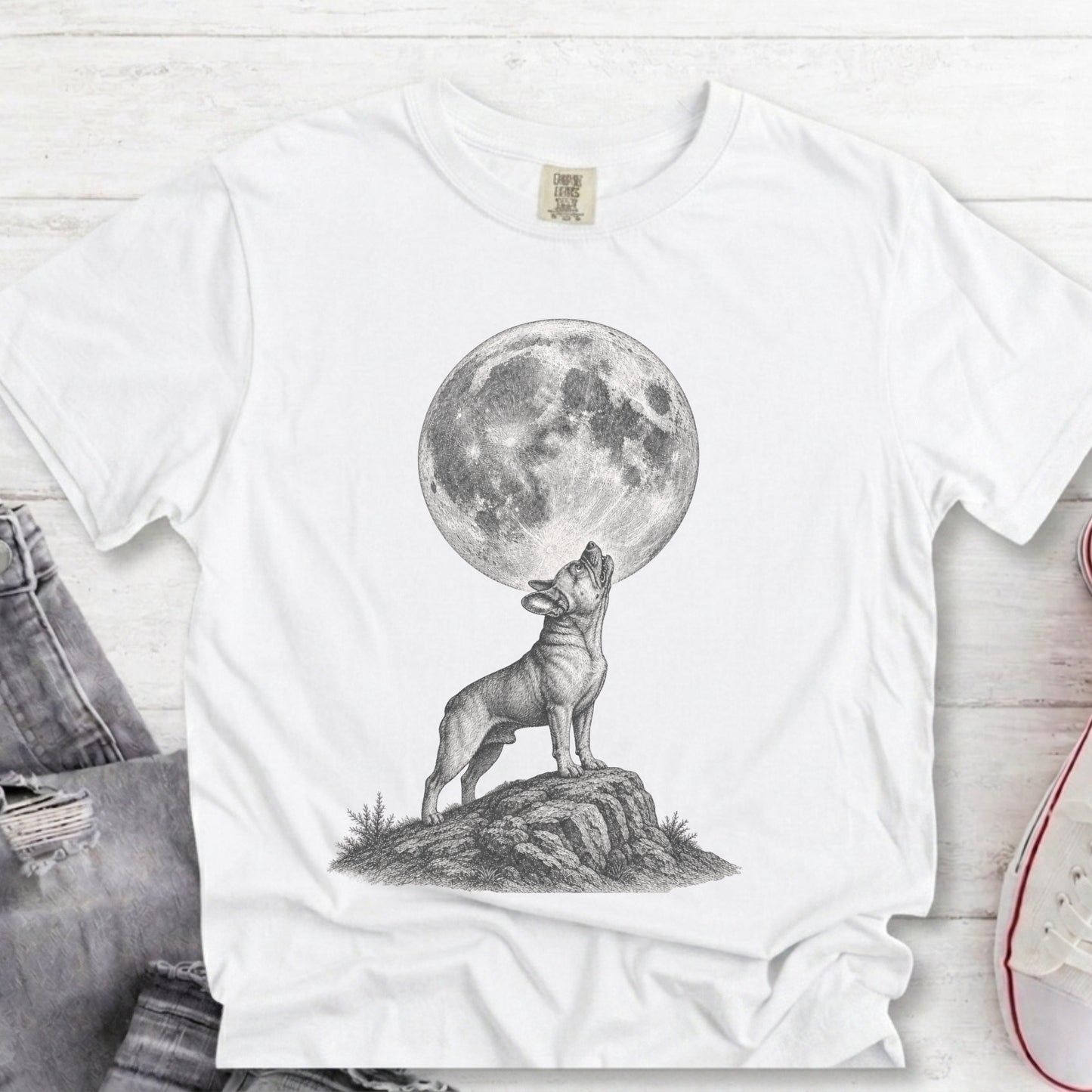 A white short-sleeve crewneck t-shirt featuring a black and white graphic of a dog howling at a large full moon. The shirt is styled in a flat-lay arrangement on a white wood background, partially overlaid with a pair of blue jeans on the left and a red and white sneaker on the right.
