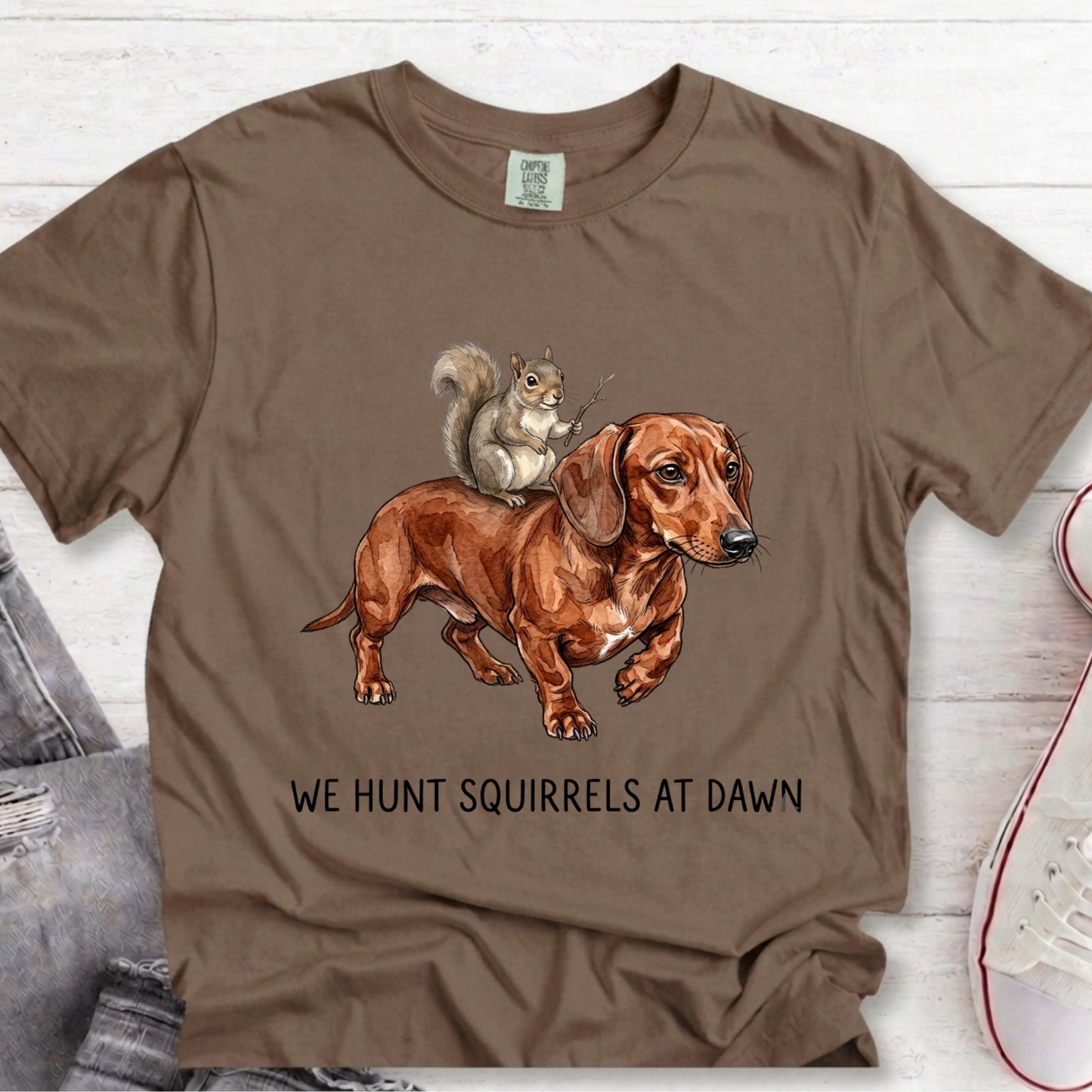 A brown short-sleeve graphic T-shirt laid flat on a light wood surface, featuring an illustration of a dachshund with a squirrel on its back and the text 'WE HUNT SQUIRRELS AT DAWN'.