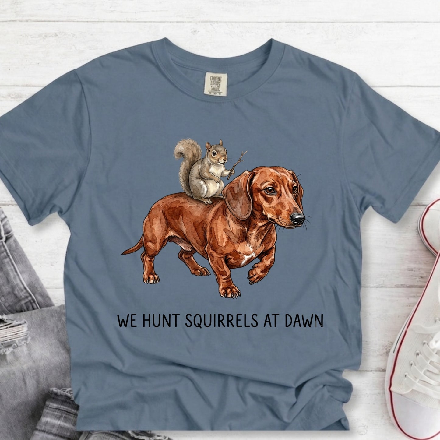 A slate blue graphic t-shirt laid flat on a white wood background, featuring an illustration of a brown dachshund with a squirrel on its back and the text 'WE HUNT SQUIRRELS AT DAWN'.