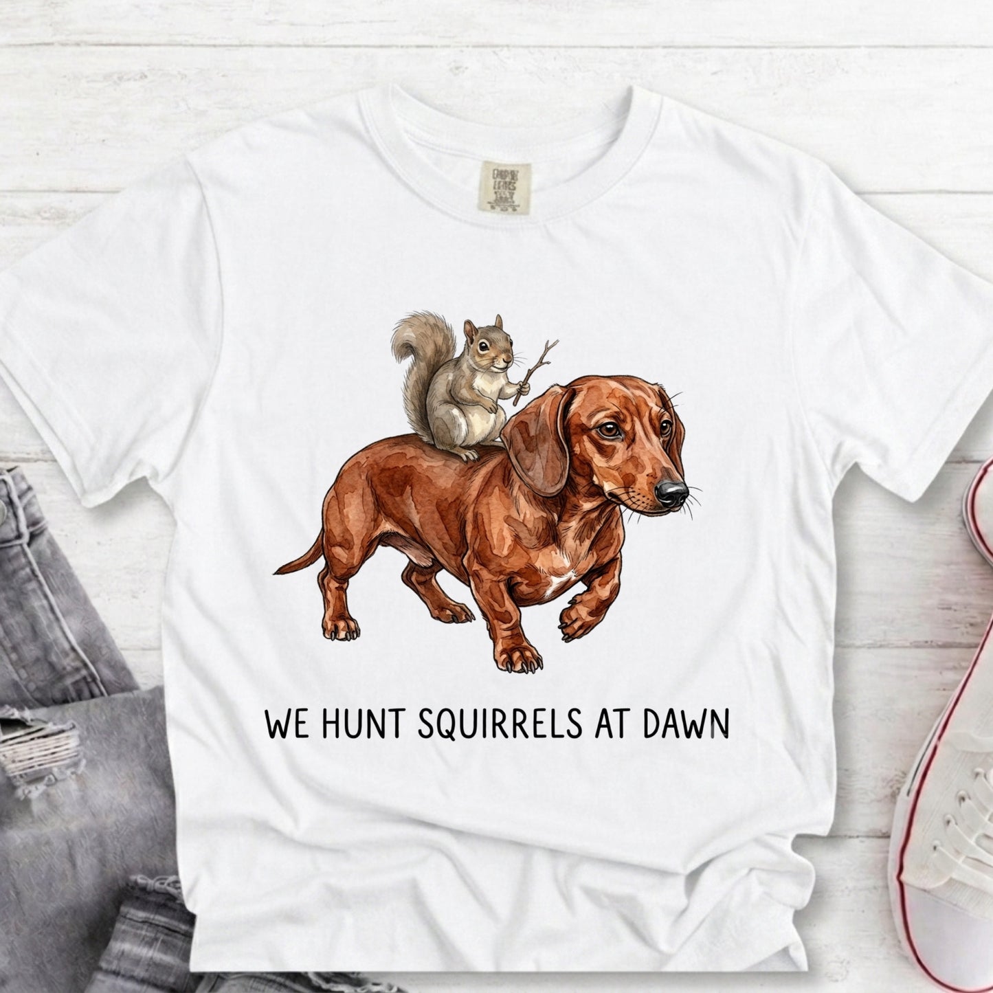 A white short-sleeve graphic T-shirt laid flat on a light wood surface, featuring an illustration of a brown dachshund with a gray squirrel on its back and the text 'WE HUNT SQUIRRELS AT DAWN' below. Partial jeans and a sneaker are visible at the edges as styling props.