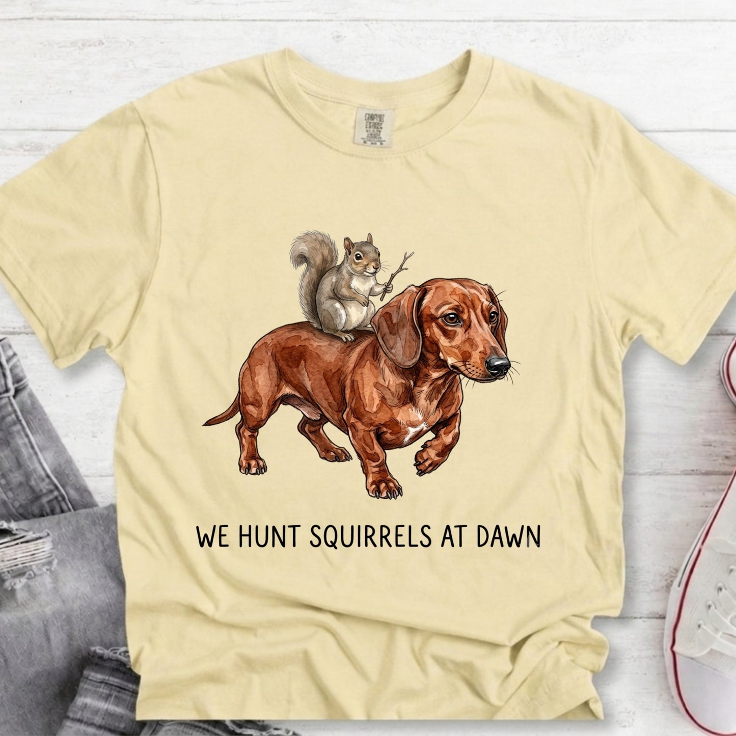 A pale yellow graphic t-shirt laid flat on a white wood background, featuring a brown dachshund and a squirrel illustration with the text 'WE HUNT SQUIRRELS AT DAWN'.