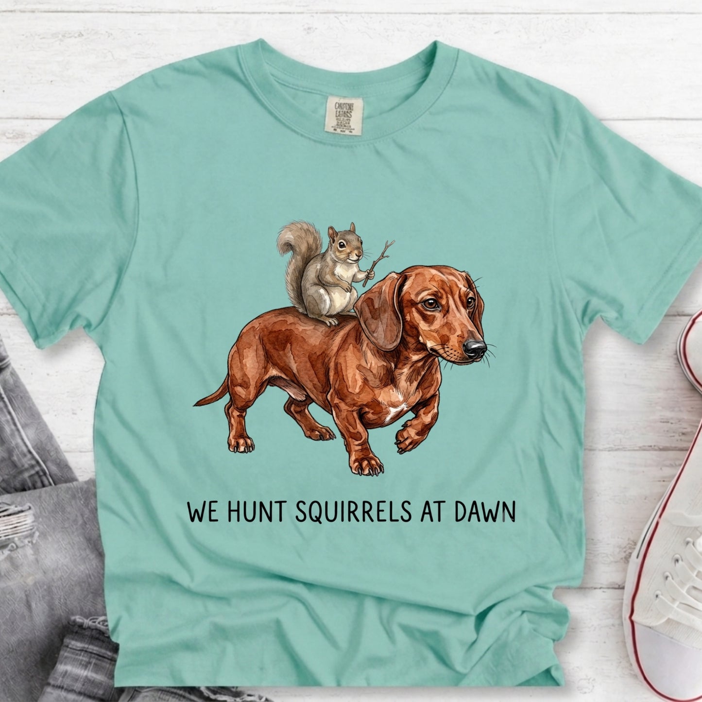 A mint green graphic t-shirt laid flat on a white wood background. The shirt features a brown dachshund and a gray squirrel illustration with the text 'WE HUNT SQUIRRELS AT DAWN' printed below.
