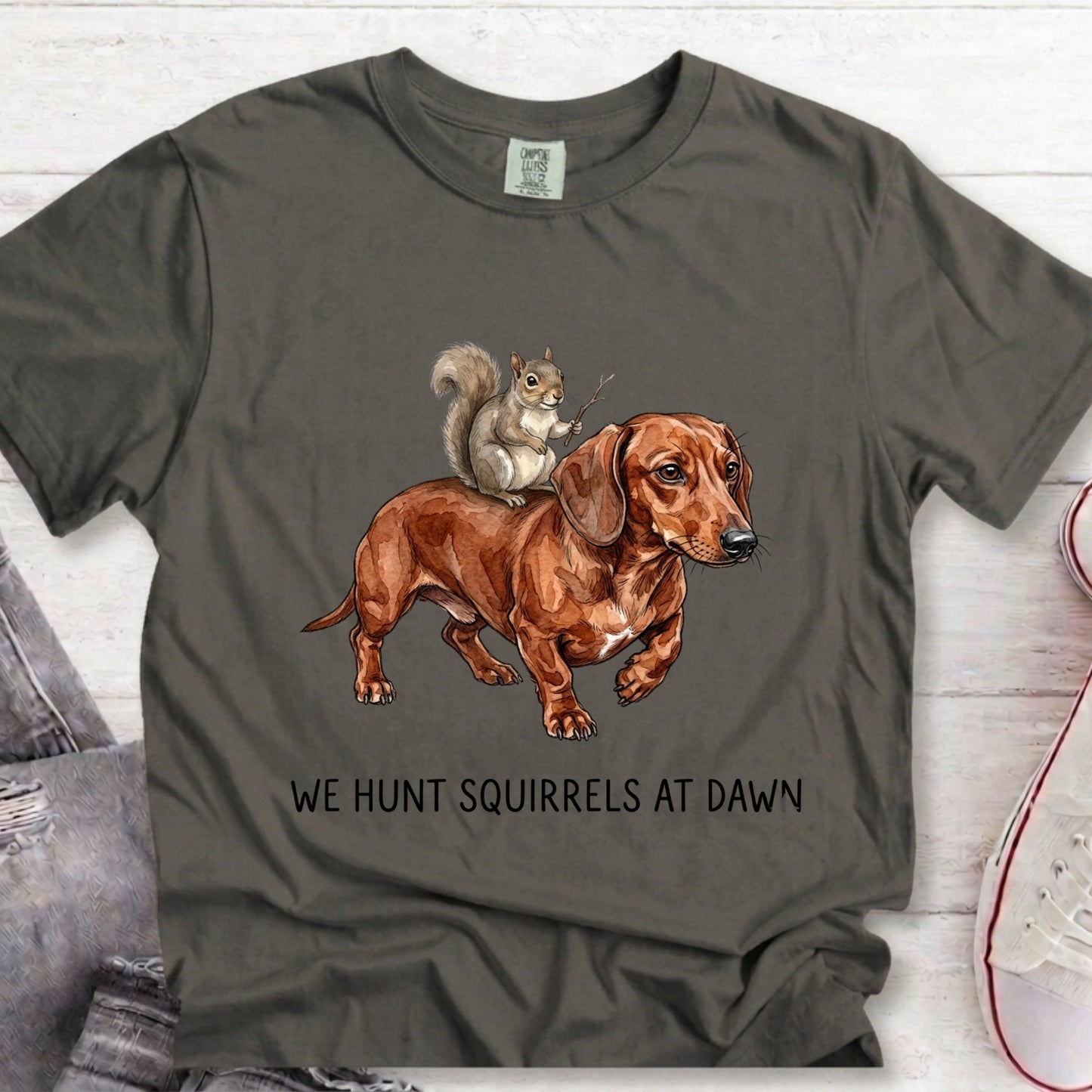 A dark gray t-shirt featuring a graphic of a brown dachshund with a squirrel on its back, with the text 'WE HUNT SQUIRRELS AT DAWN' printed below. The shirt is displayed flat on a light wood background with jeans and a sneaker partially visible.