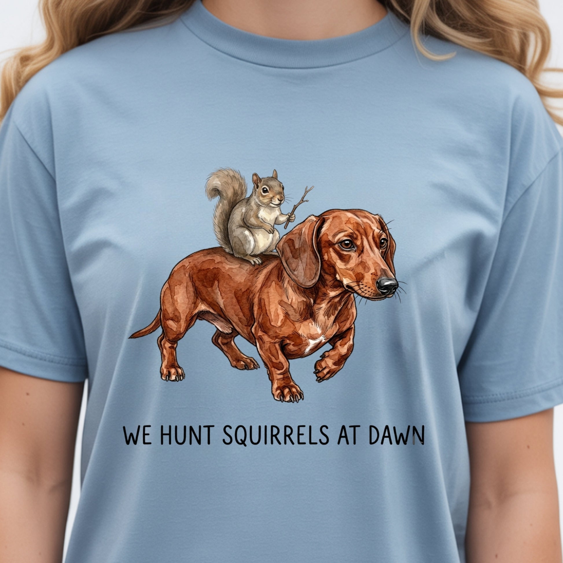 A person wearing a light blue crewneck t-shirt with a graphic of a brown dachshund and a gray squirrel, with the text 'WE HUNT SQUIRRELS AT DAWN' printed below.