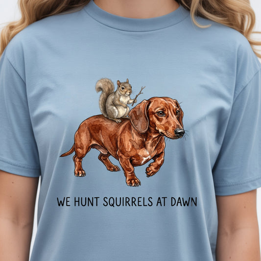 A person wearing a light blue crewneck t-shirt with a graphic of a brown dachshund and a gray squirrel, with the text 'WE HUNT SQUIRRELS AT DAWN' printed below.