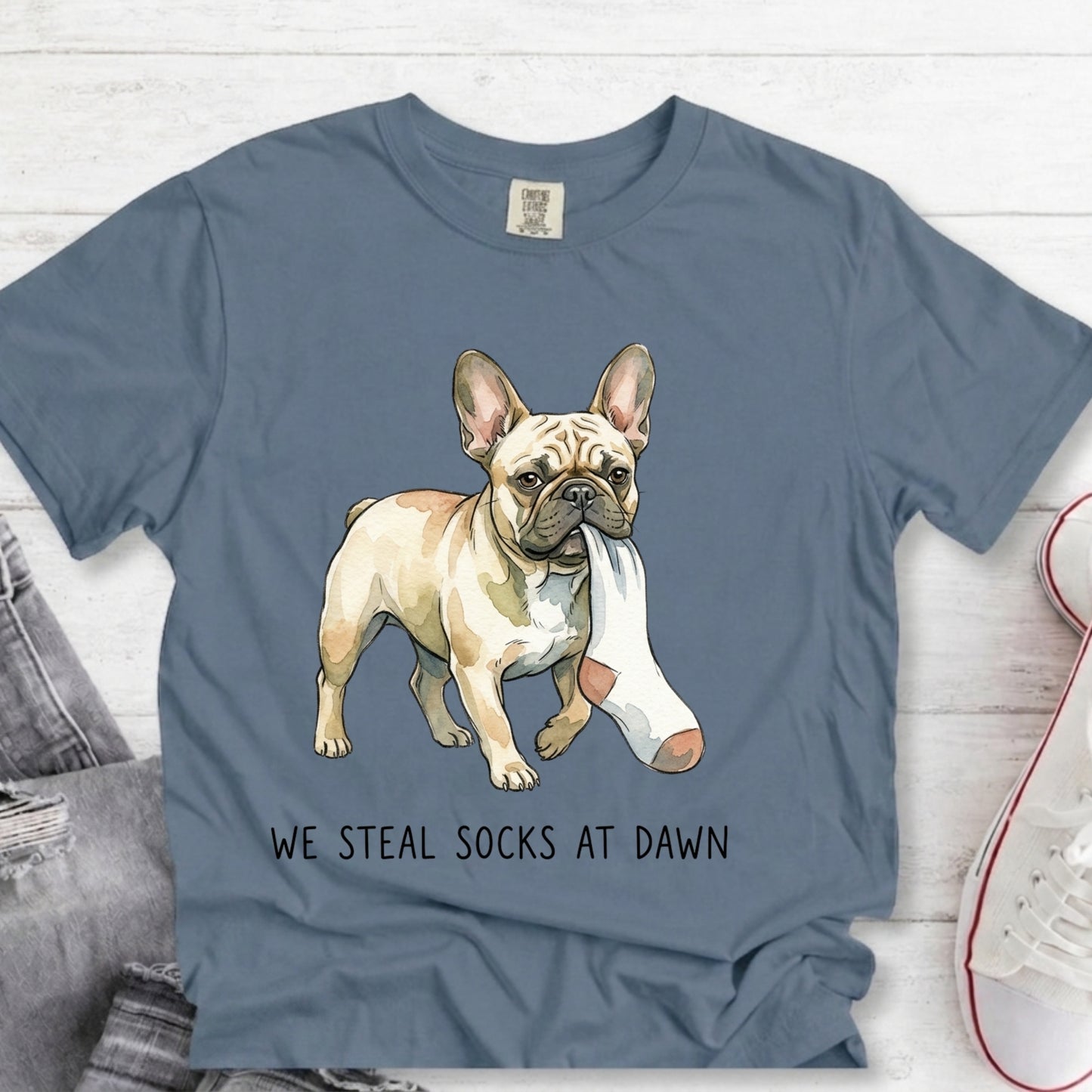 A slate blue graphic t-shirt featuring a French Bulldog carrying a white sock, with the text 'WE STEAL SOCKS AT DAWN' printed below. The t-shirt is styled in a flat-lay arrangement on a white wood background with jeans and a sneaker partially visible.
