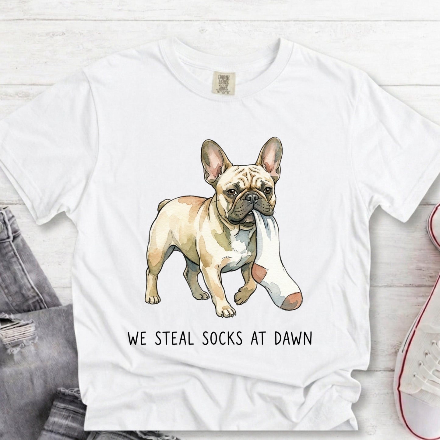 A white short-sleeve graphic T-shirt laid flat on a light wood surface, featuring an illustrated French bulldog wearing a pink-and-white sock and the text 'WE STEAL SOCKS AT DAWN.' A folded pair of jeans and part of a sneaker are visible as styling props.