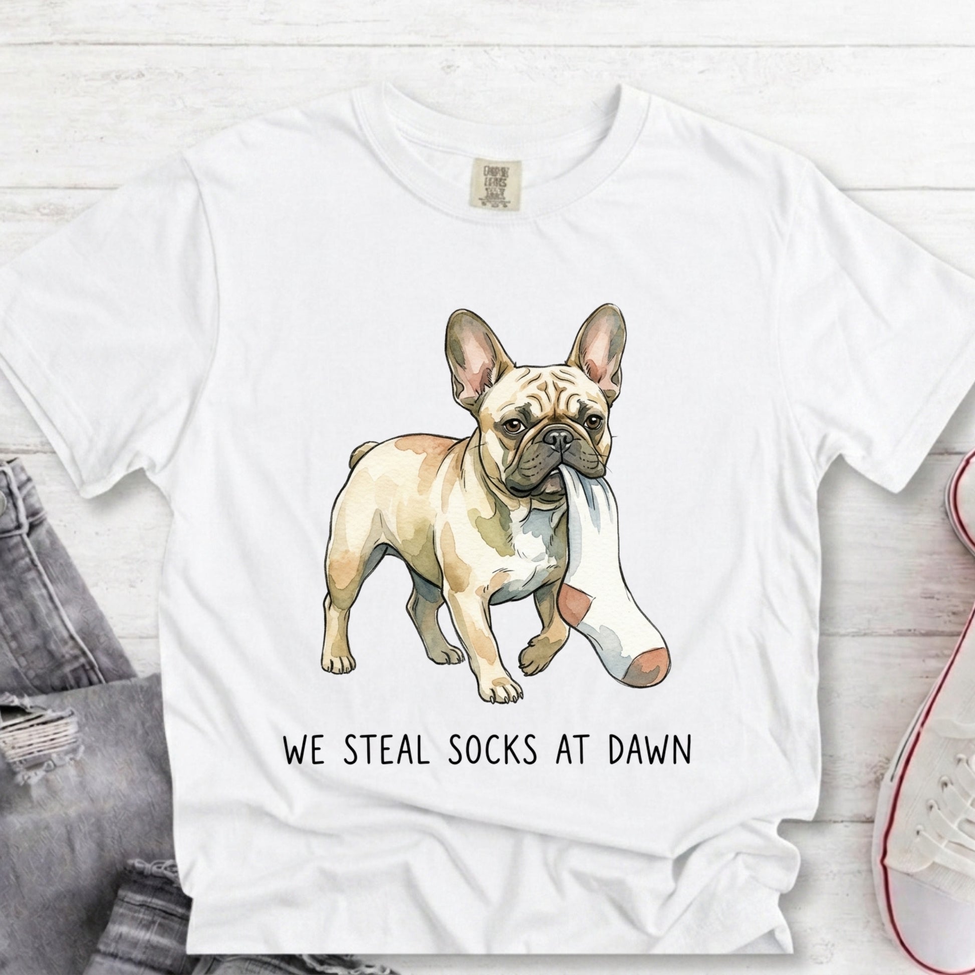 A white short-sleeve graphic T-shirt laid flat on a light wood surface, featuring an illustrated French bulldog wearing a pink-and-white sock and the text 'WE STEAL SOCKS AT DAWN.' A folded pair of jeans and part of a sneaker are visible as styling props.