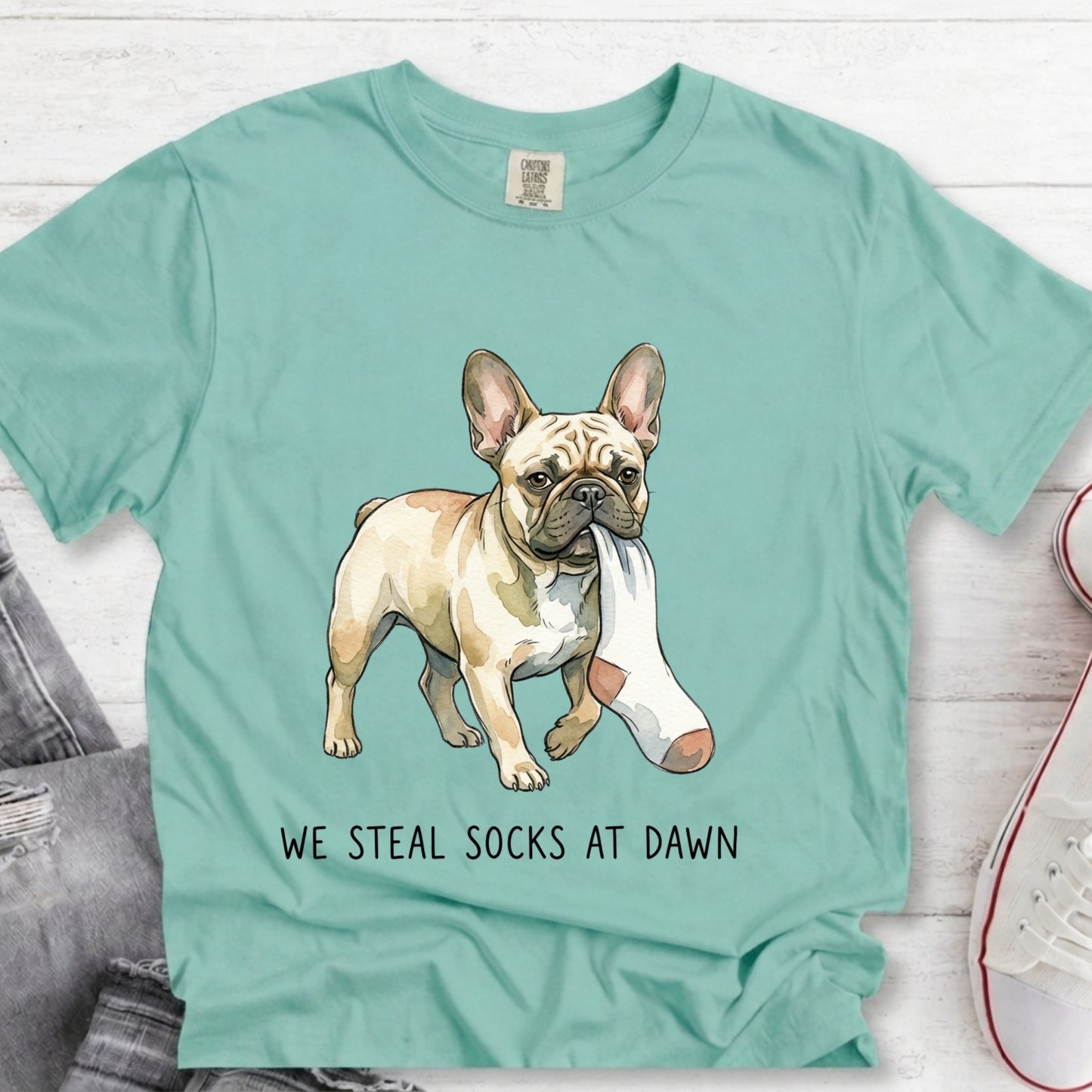 A mint green graphic t-shirt laid flat on a white wood background, featuring an illustration of a French Bulldog holding a white sock in its mouth, with the text 'WE STEAL SOCKS AT DAWN' printed below.