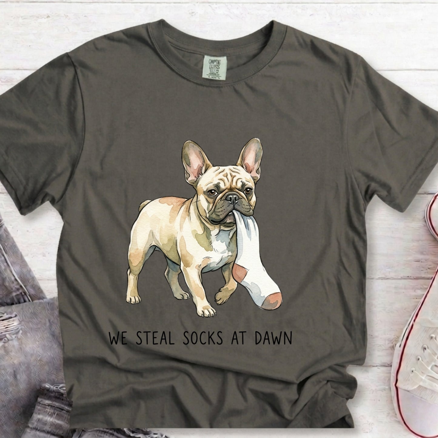A dark gray t-shirt featuring a graphic of a French Bulldog wearing a white cast on one leg, with the text 'WE STEAL SOCKS AT DAWN' printed below. The t-shirt is styled in a flat-lay arrangement on a white wood background with jeans and a sneaker partially visible.