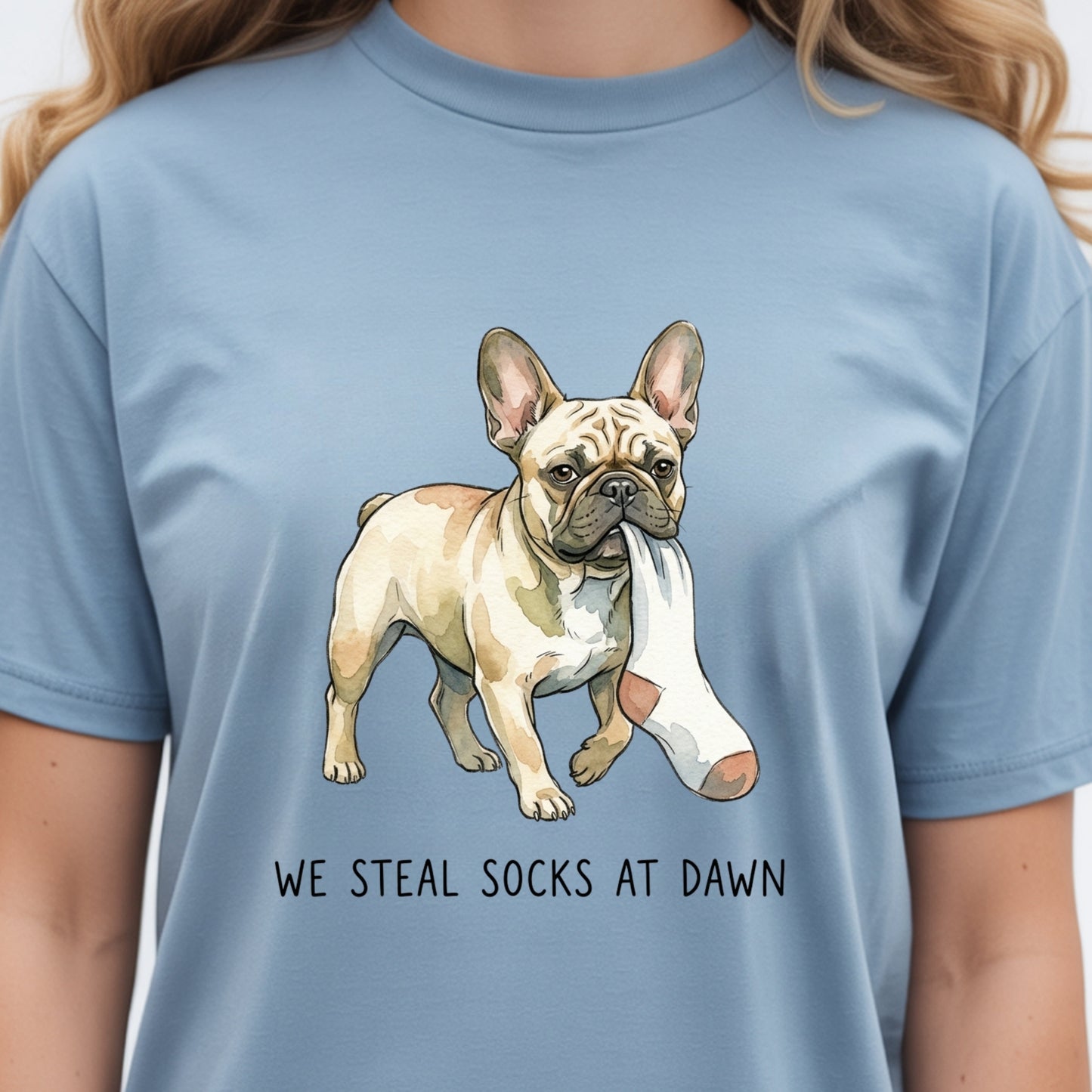 A light blue crewneck t-shirt featuring a graphic of a French Bulldog carrying a white cast on its back, with the text 'WE STEAL SOCKS AT DAWN' printed below. The shirt is shown on a model.