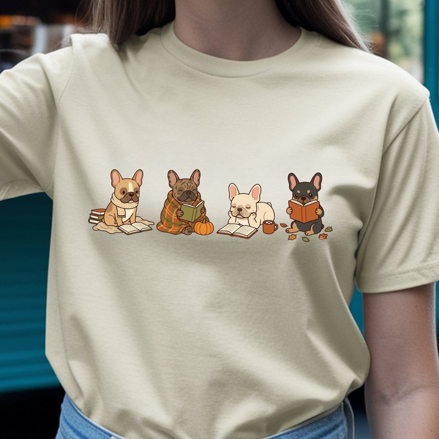 A person wearing a light beige crewneck t-shirt with a graphic print of four French bulldogs reading books on the chest, paired with blue jeans.