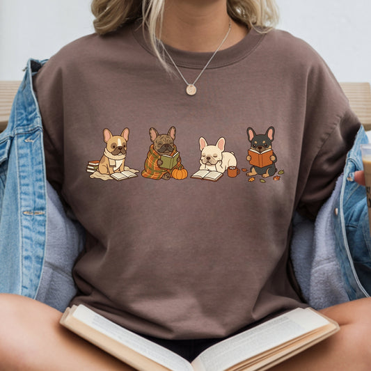 A person wearing a brown crewneck t-shirt with a graphic of four French bulldogs reading books. The person is also wearing an open blue denim jacket and holding an open book.