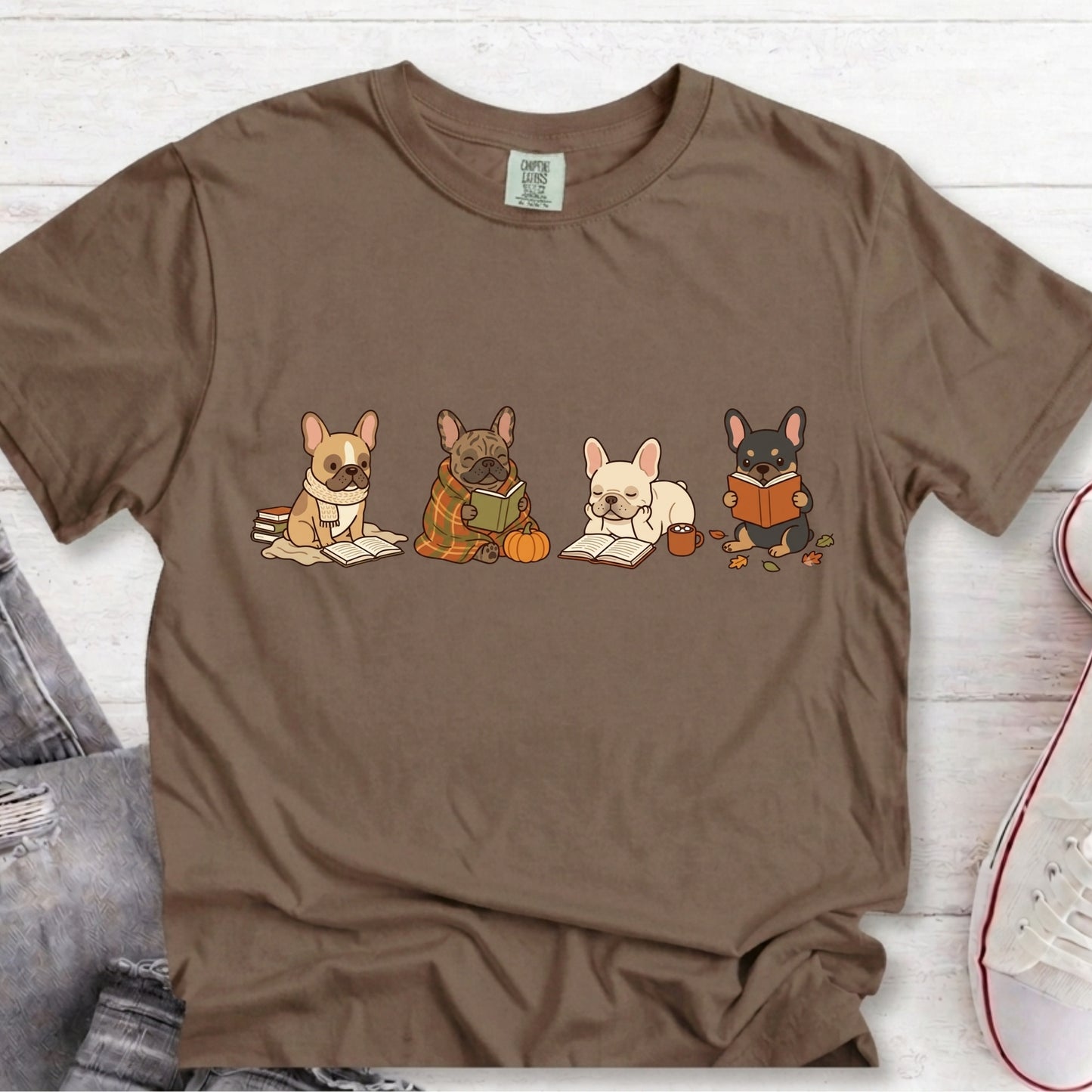 A brown short-sleeve crewneck t-shirt featuring a graphic of four cute French Bulldog characters reading books, laid flat on a white wood background.